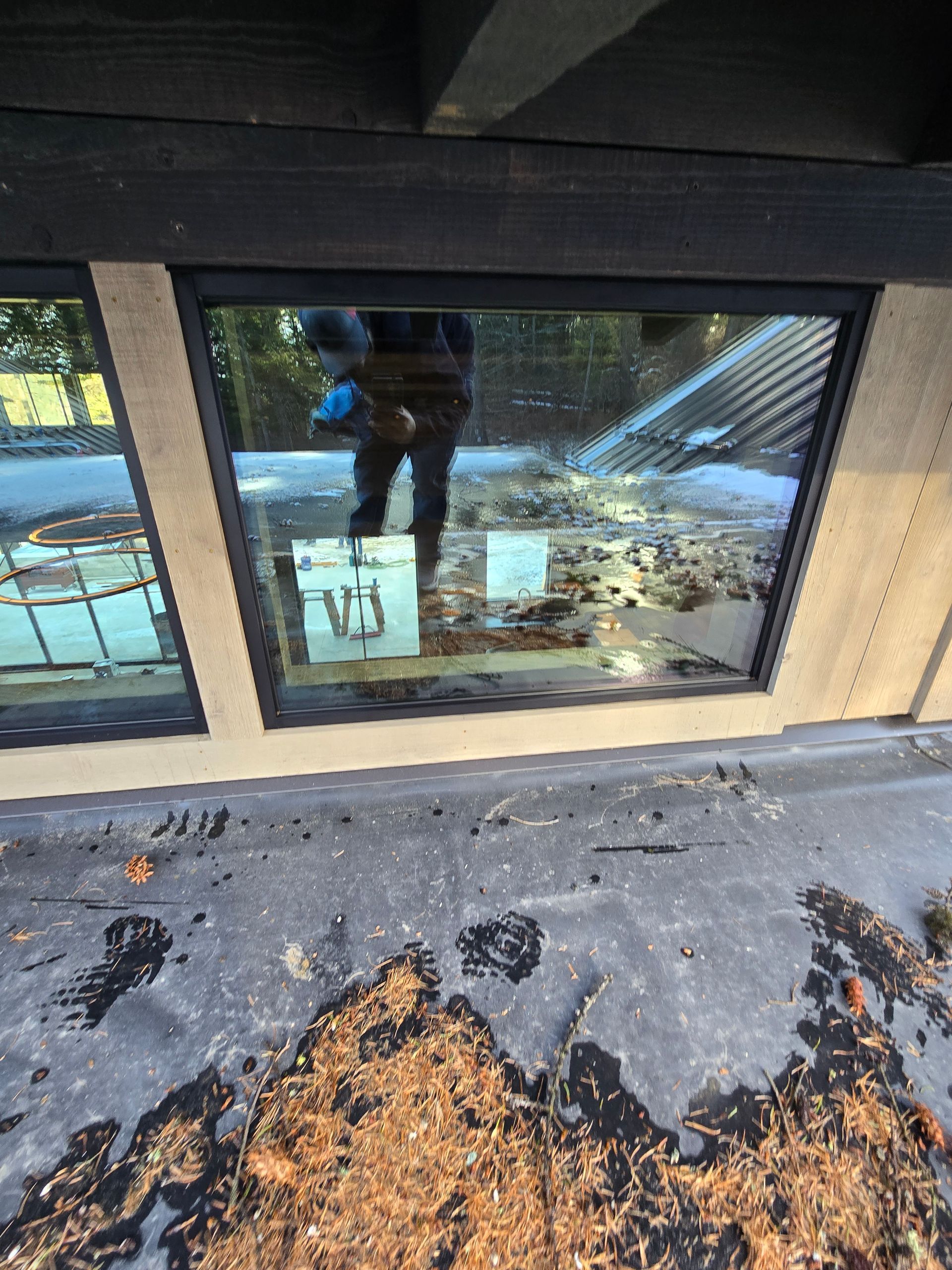 A view through a window onto a flat, dark, debris-covered roof, reflecting a person taking the photo.