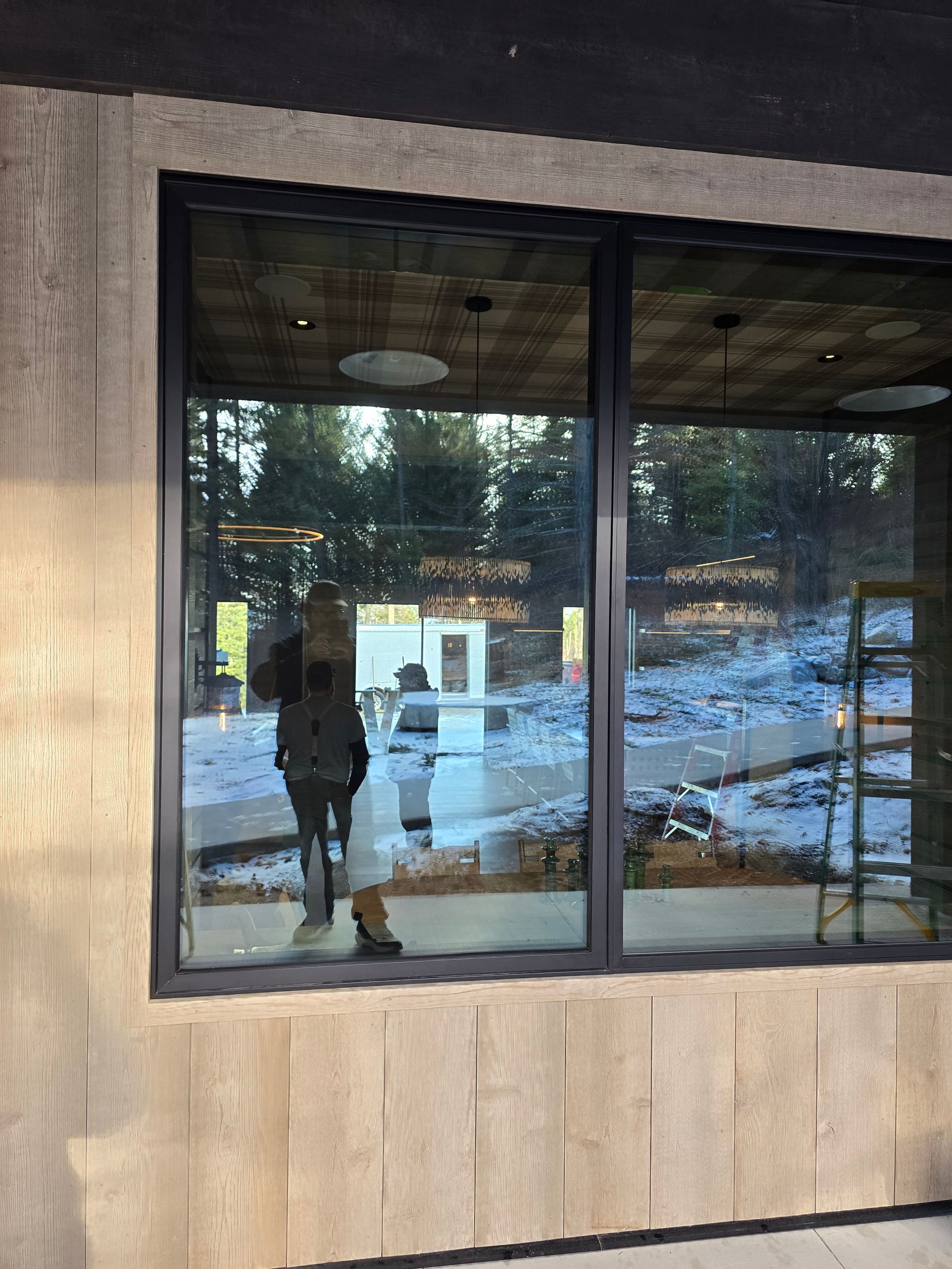 A person reflected in the large window of a modern building under construction with wood paneling and snow outside.