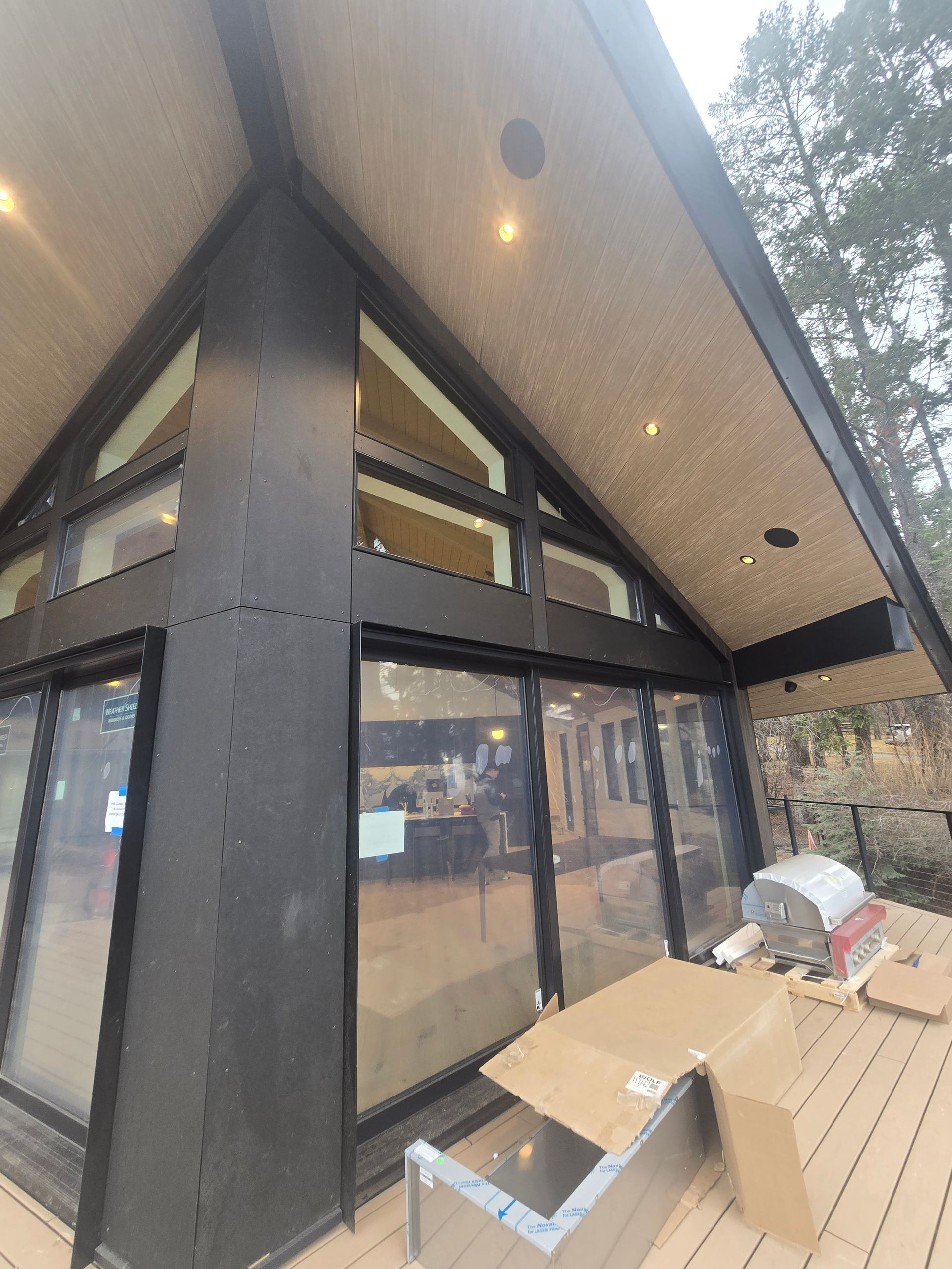 A low-angle view of a modern A-frame cabin exterior with black metal framing, glass windows, and a wood-paneled deck.