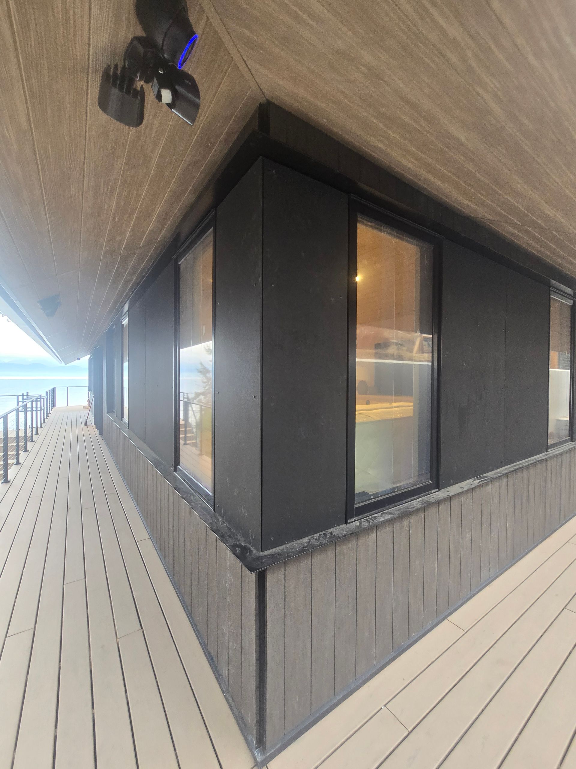A low-angle view of a building corner with black exterior walls, large windows, and a slatted deck overlooking water.