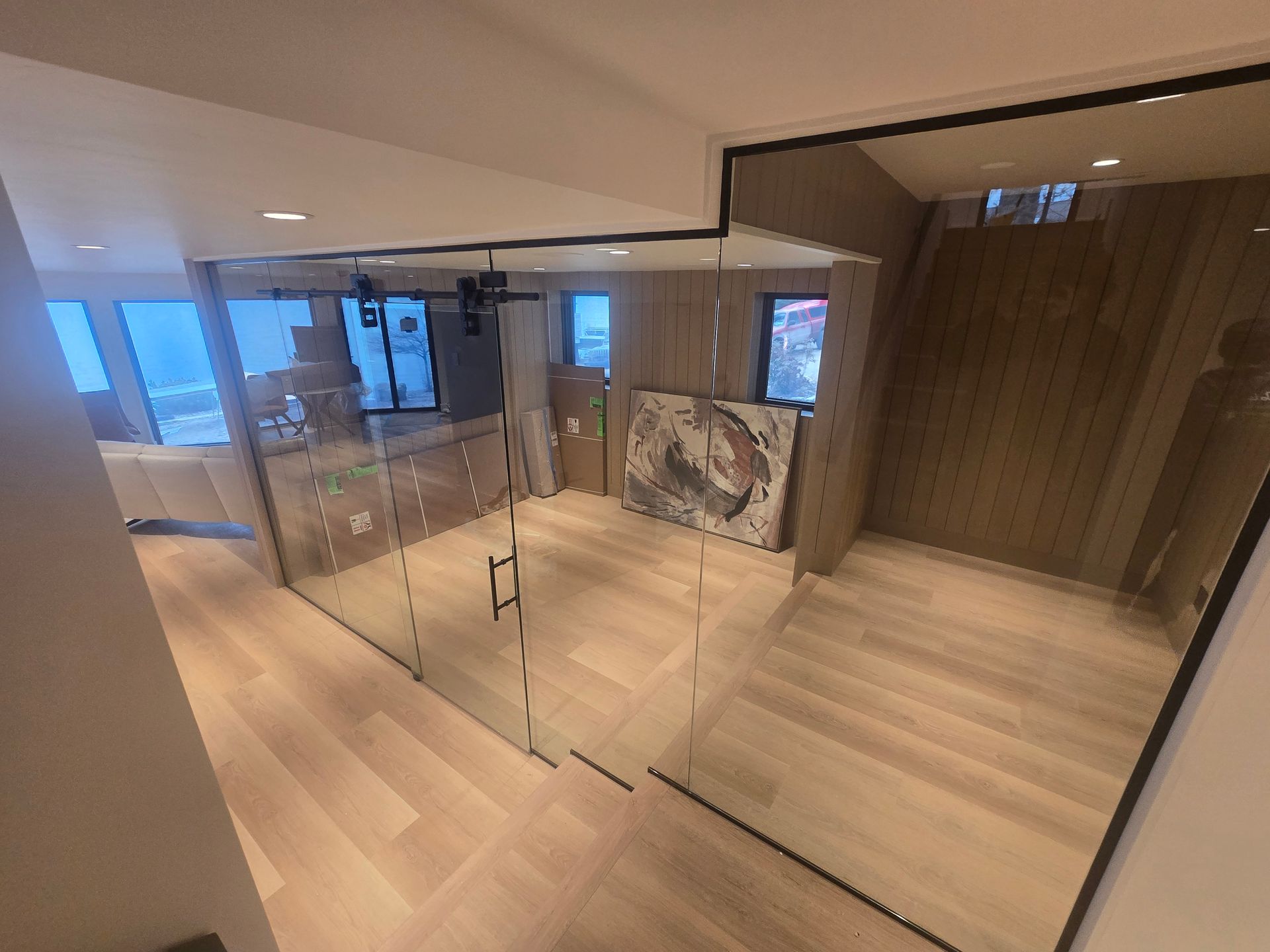 A modern interior view of a room with light wood floors, a glass-enclosed office space, and a piece of abstract art.
