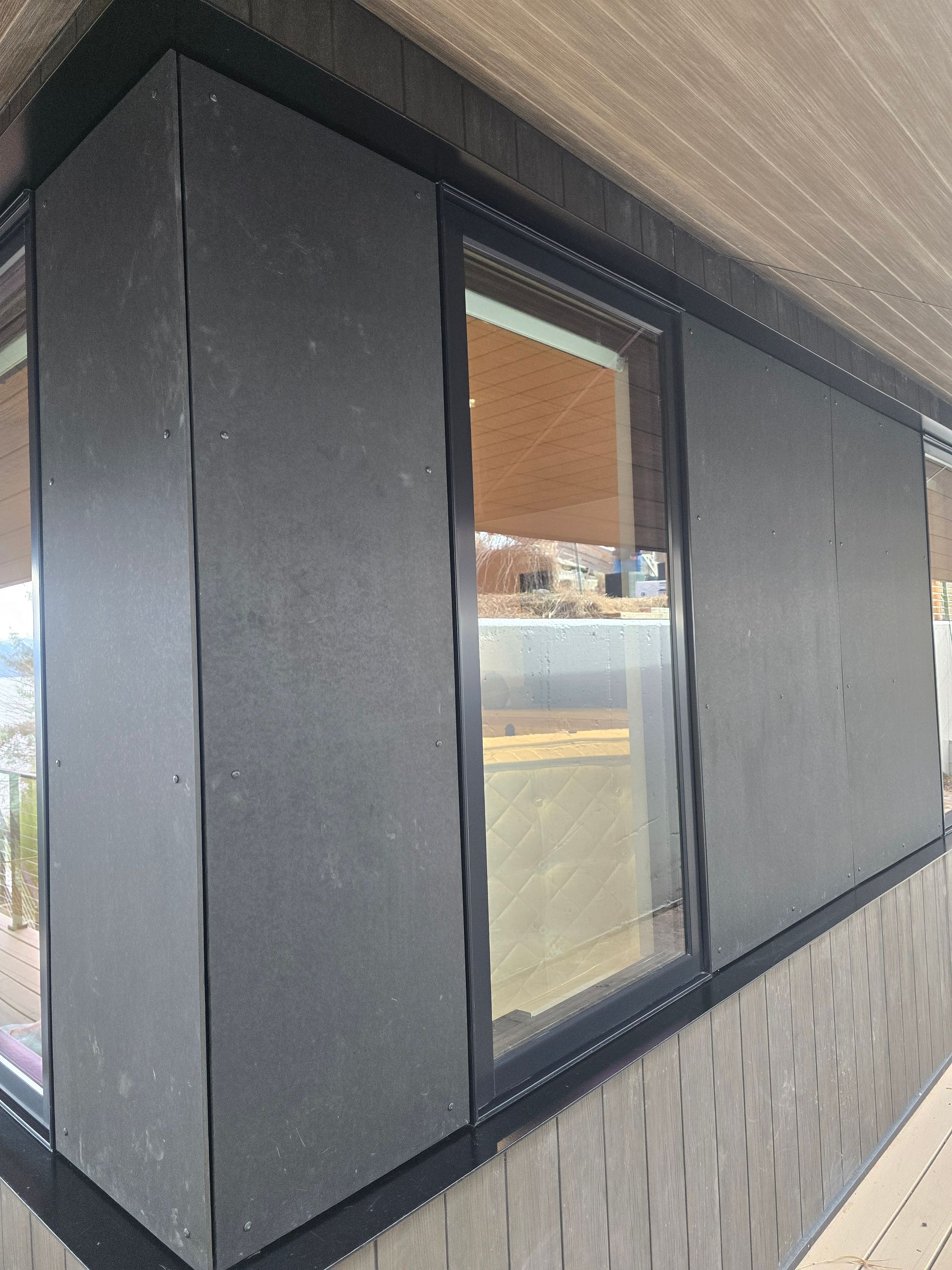 A close-up view of a building exterior featuring grey paneling, a large window, and black trim against a wooden ceiling.