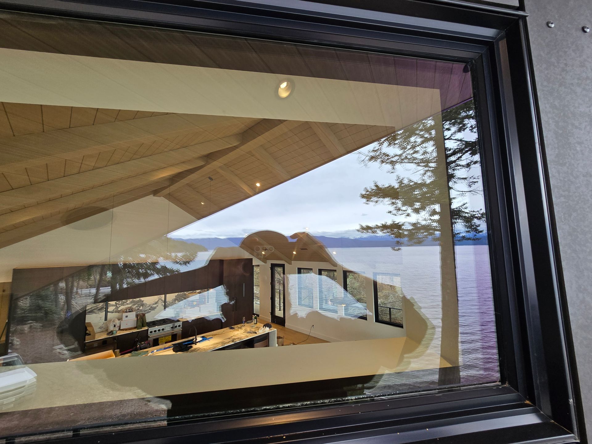 A window reflects an indoor room with a high wooden ceiling and a table, overlooking a lake and trees outside.