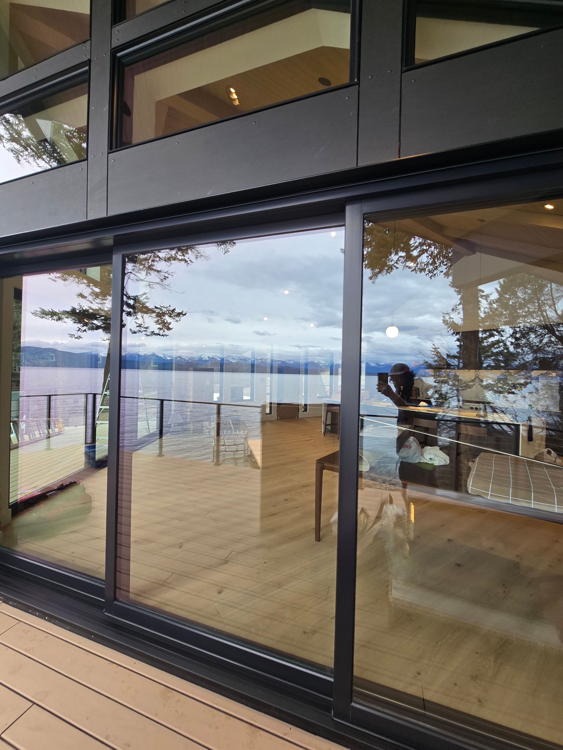 Large sliding glass doors look out onto a wooden deck, a lake, and a cloudy sky, with interior furniture reflected in glass.