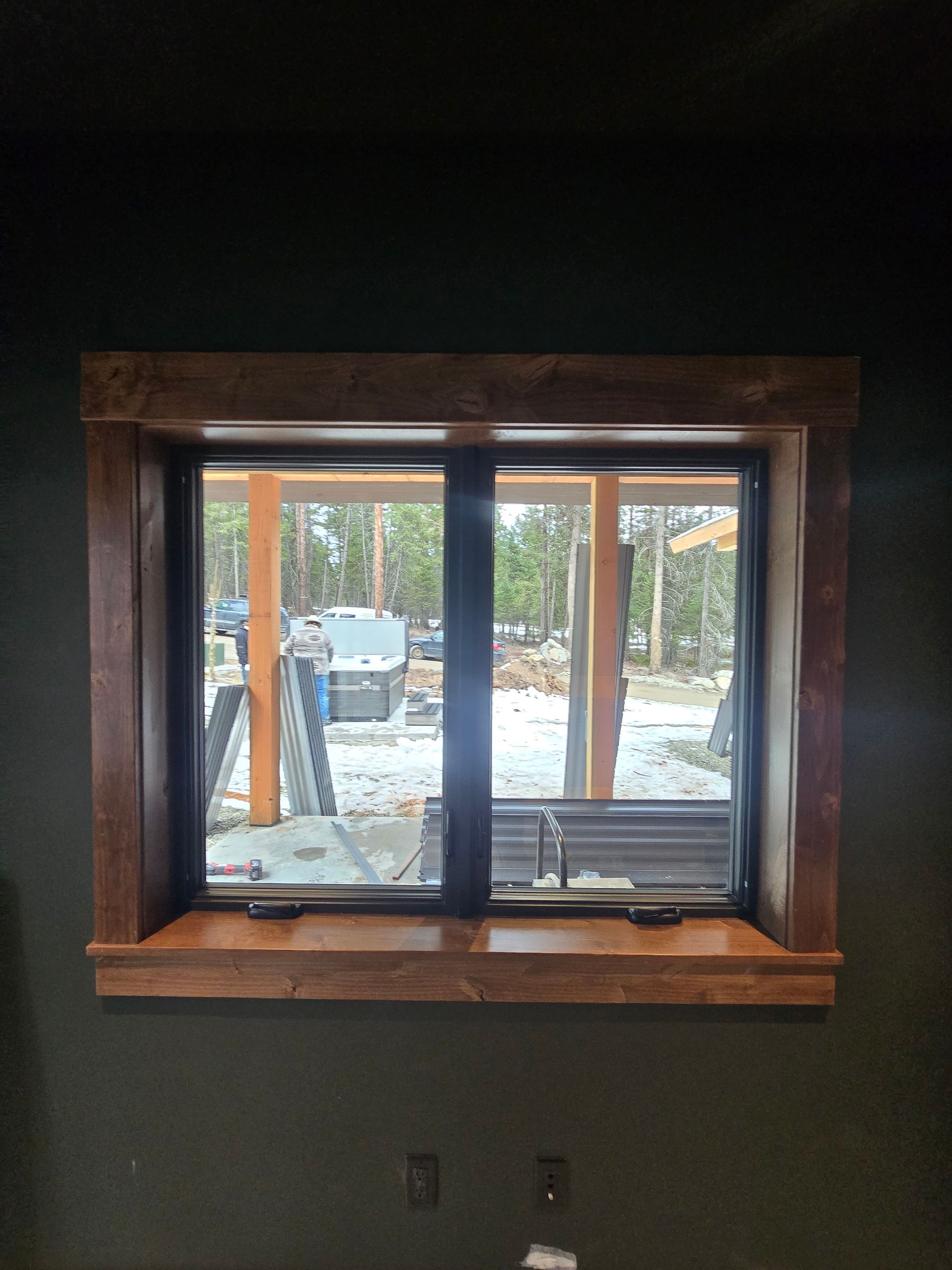 A window with thick, dark-stained wooden trim centered on a dark, textured wall, looking out onto a snowy, wooded yard.