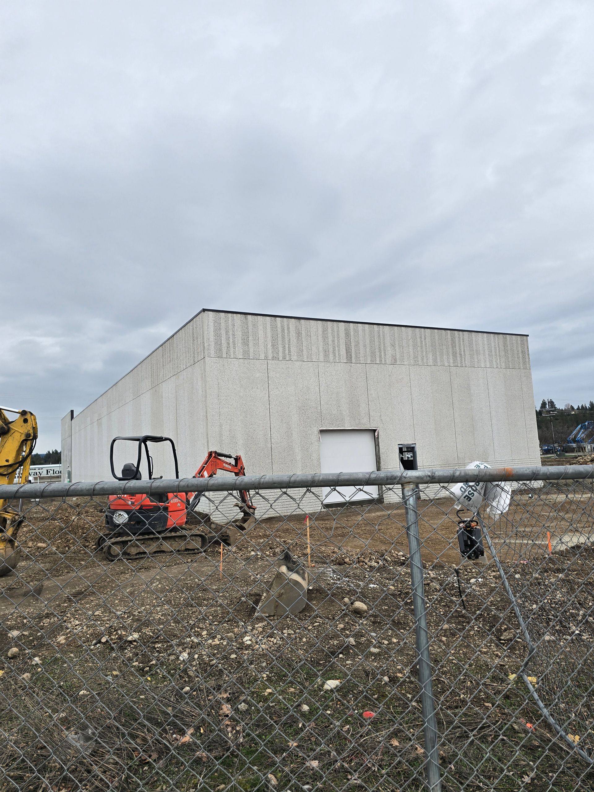 An orange mini excavator sits on a dirt construction site in front of a large, white windowless building.