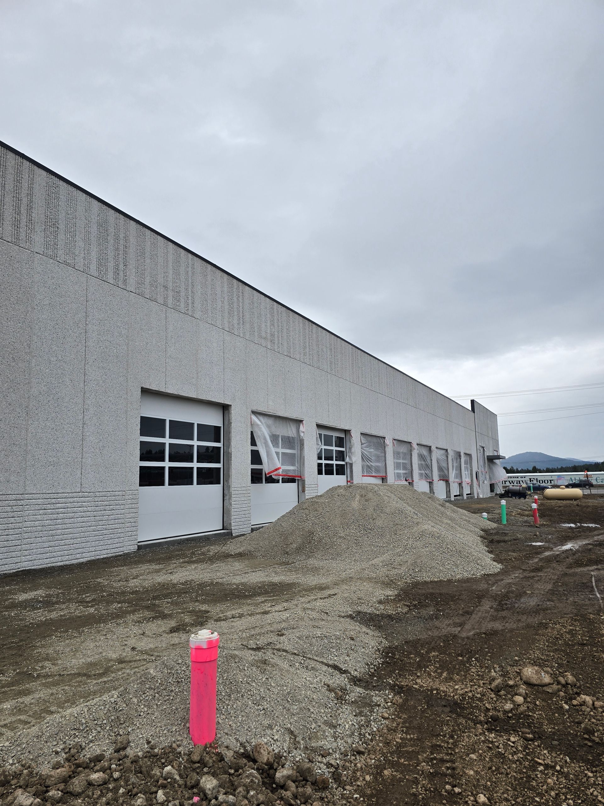 A long, textured white building with several garage doors under a cloudy sky, with a pile of gravel in the foreground.