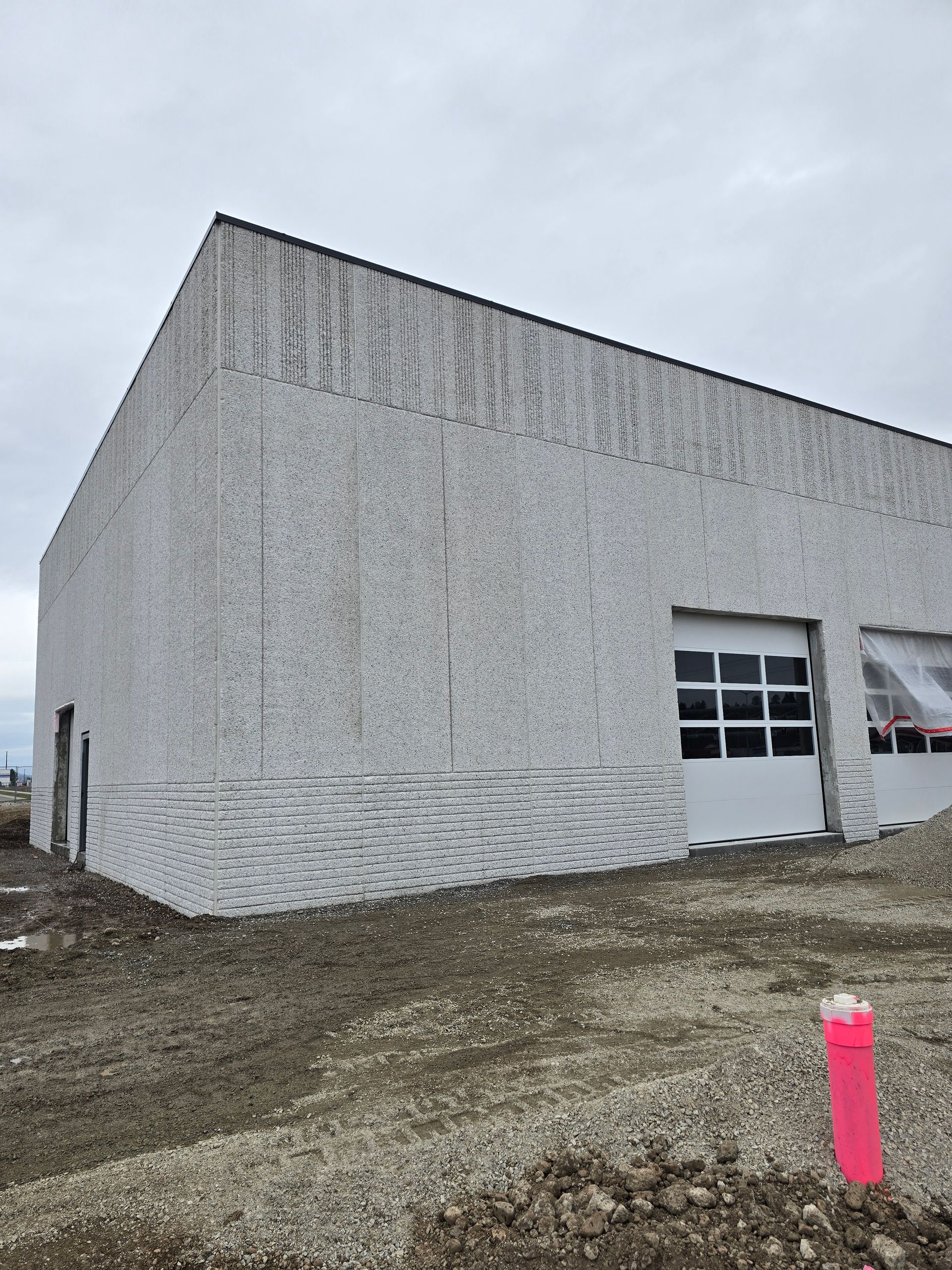 A white, textured concrete building under a cloudy sky, featuring two garage-style doors and rough ground in the foreground.