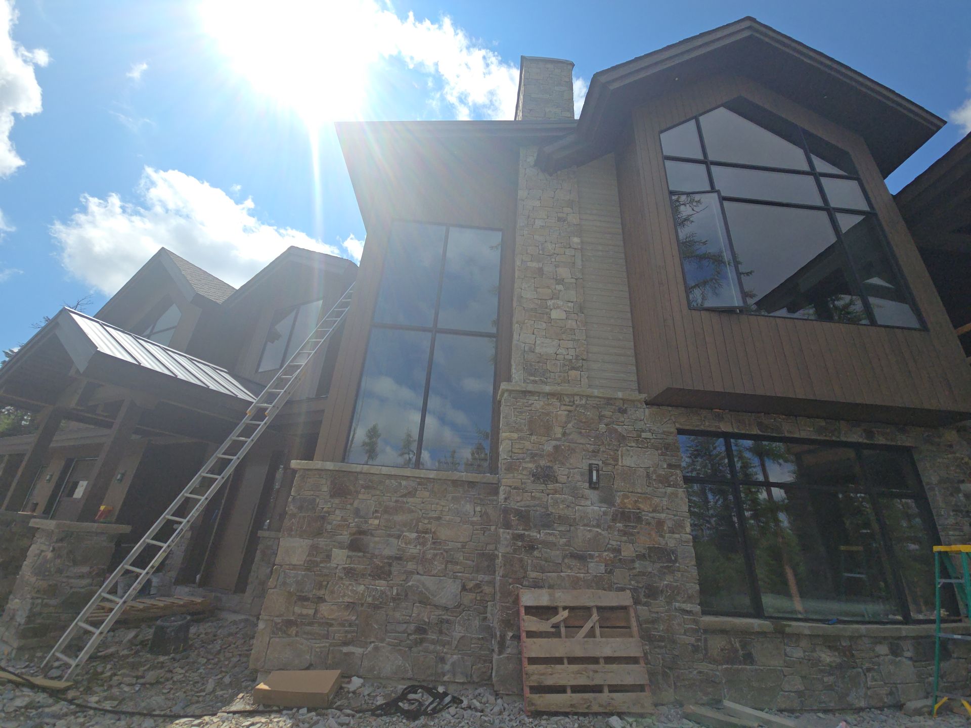 A two-story stone and wood house under construction on a sunny day with a ladder leaning against the exterior.