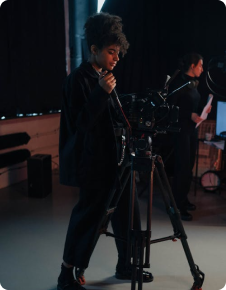Person adjusting a camera on a tripod in a dim studio, with another person in the background.