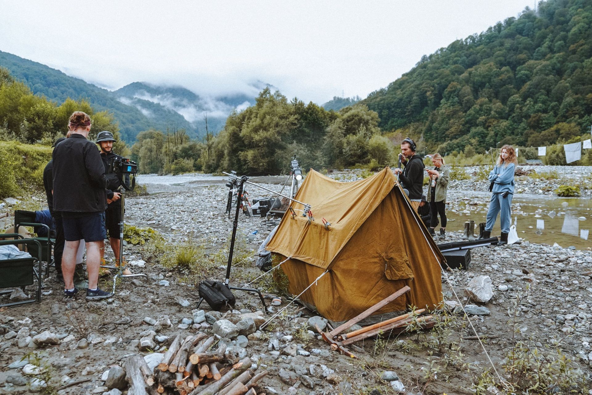 Campers surveying flood debris beside a tan tent in a rocky river valley with forested hills.