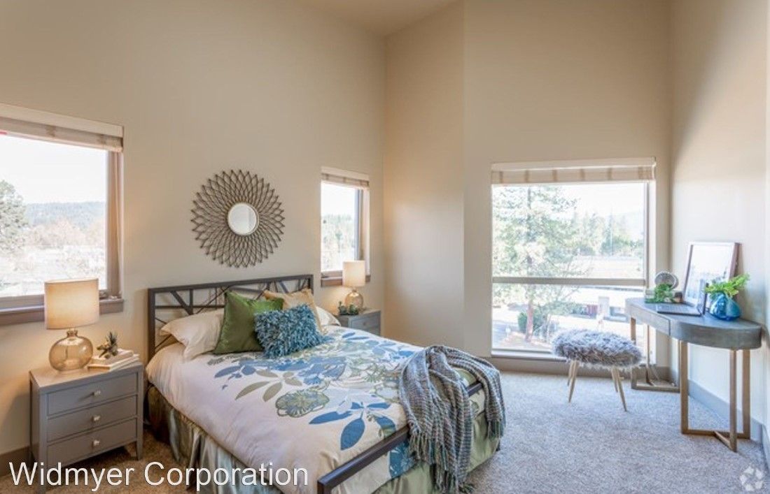 A bedroom with a bed , nightstand , desk and two windows.