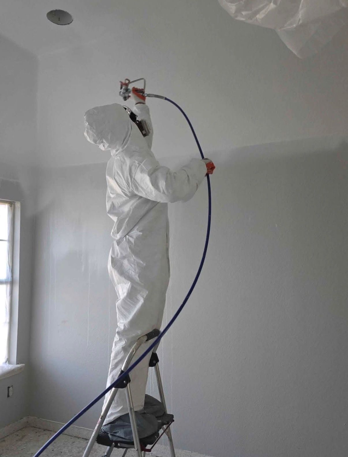 Person in protective suit spraying paint on a wall with a sprayer, standing on a ladder.
