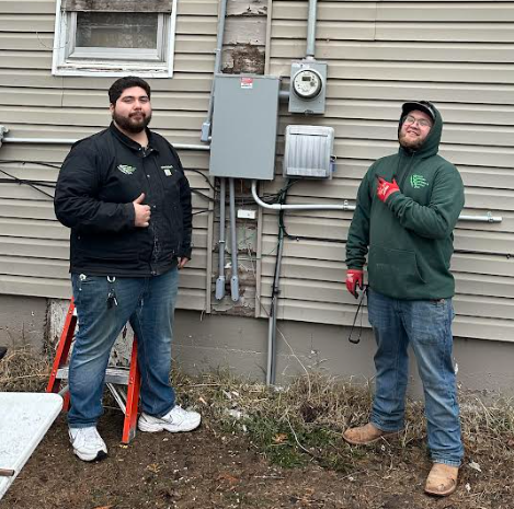 Two people stand outside next to a newly installed electrical panel and utility meter on the side of a house.