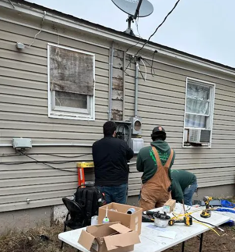 Three workers install electrical equipment on the exterior wall of a house near a table with tools.