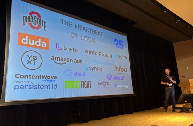A man stands in front of a large screen that says the heartbeat of local