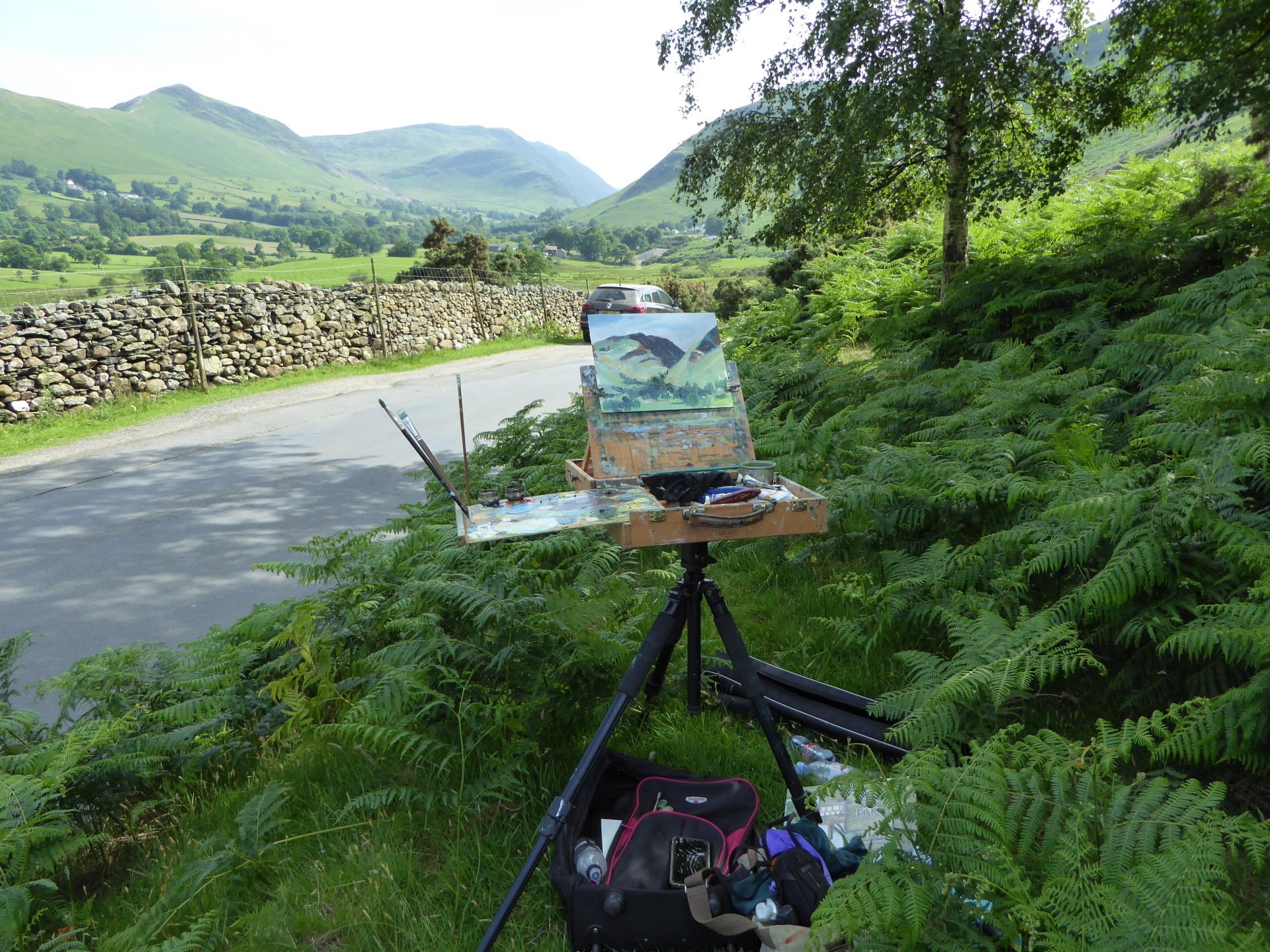An easel with a painting on it is sitting in the grass near a road.
