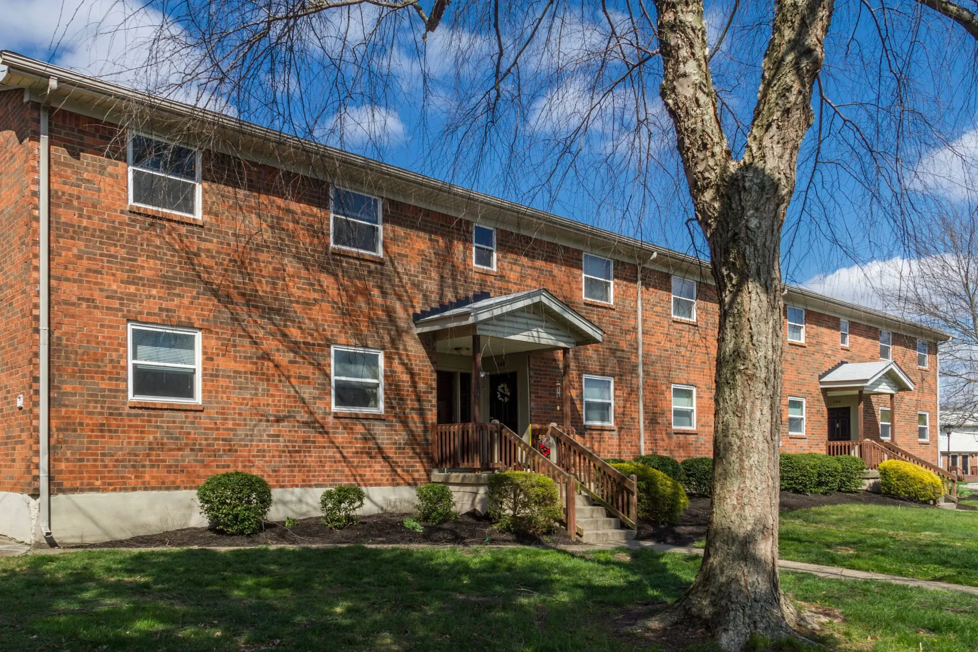 Two-story brick apartment building with trees and blue sky.