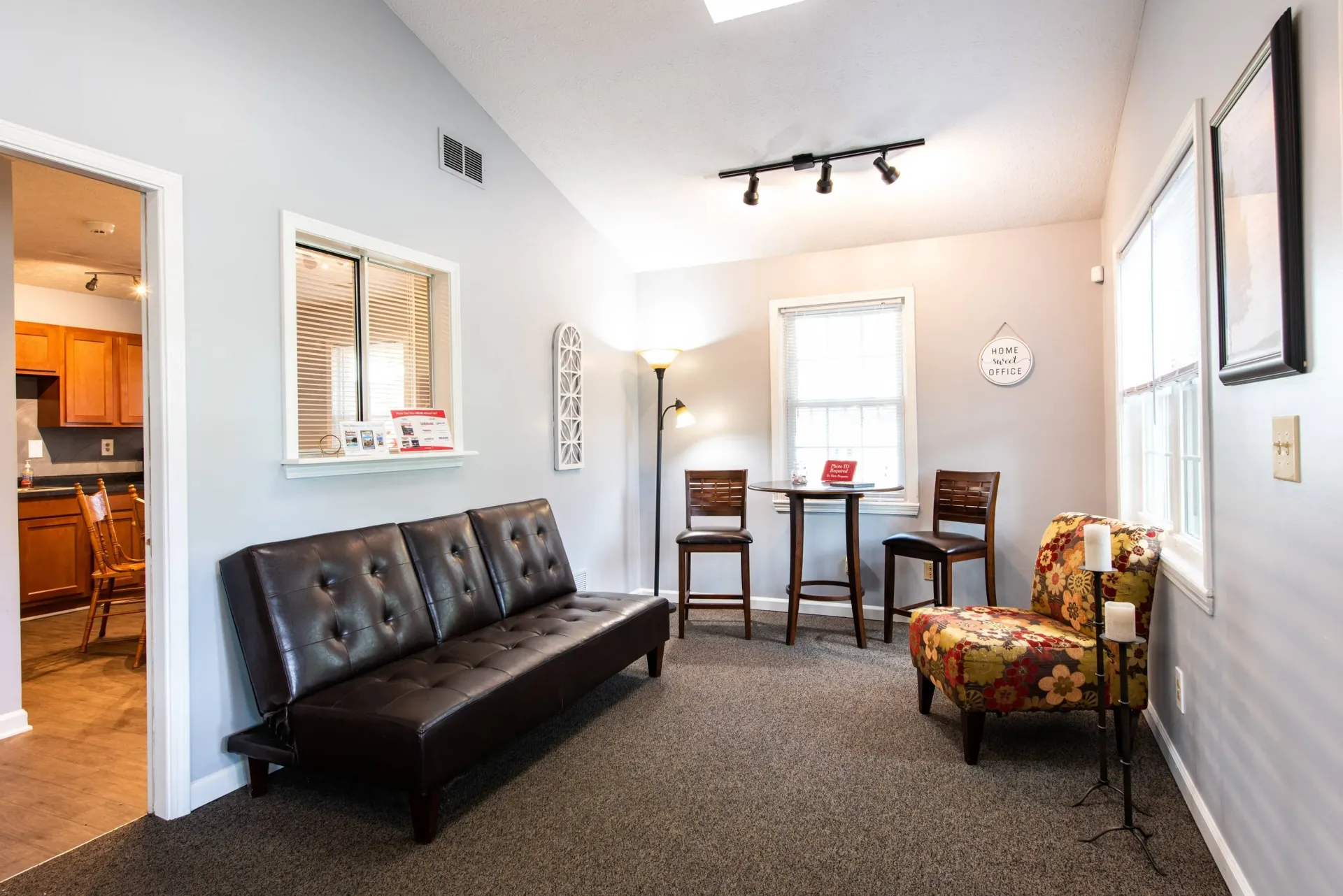 Living room with brown sofa, floral chair, round table, and two chairs; view of kitchen.