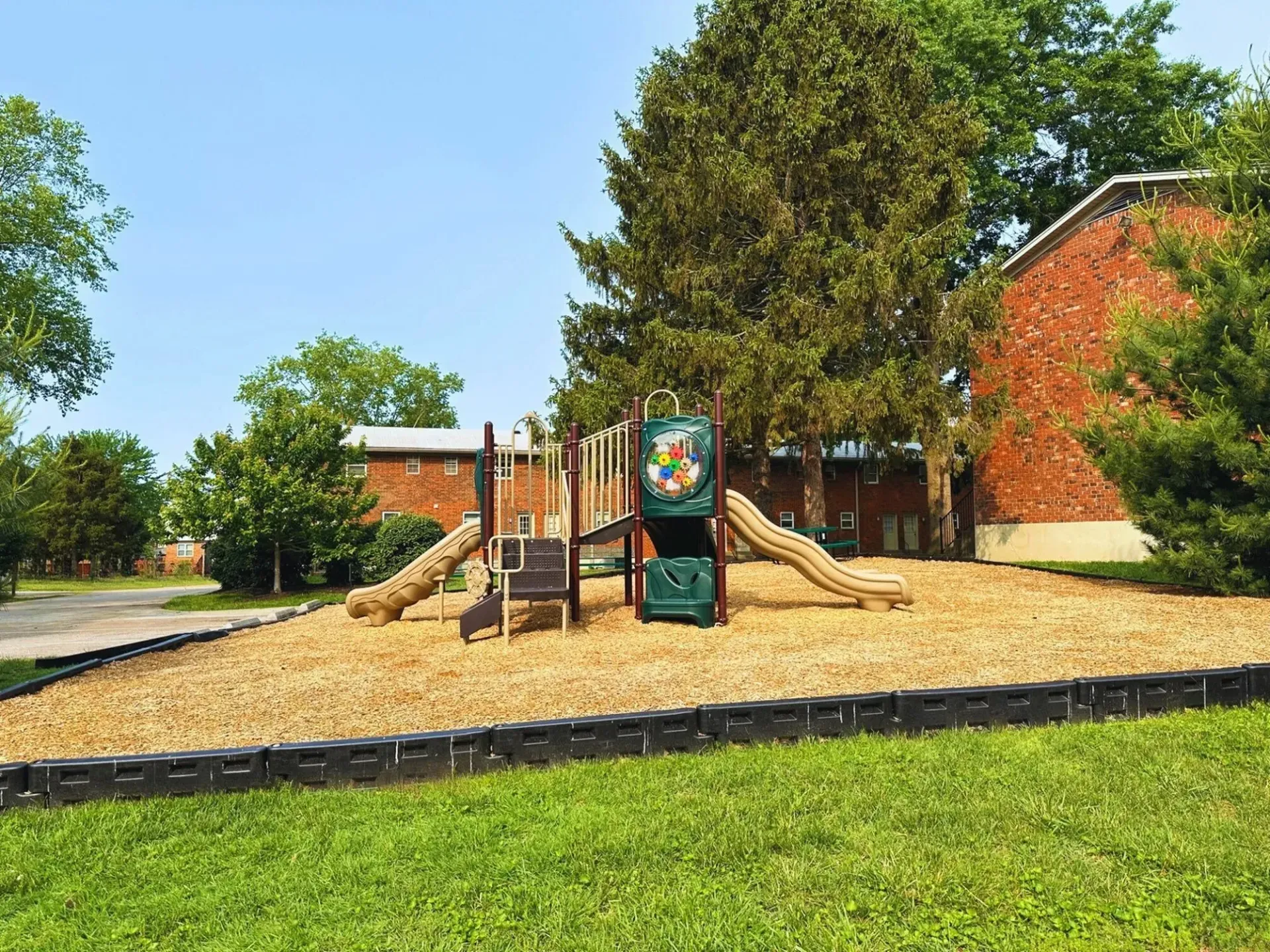 Playground with wood chips, slides, and brick buildings in the background.