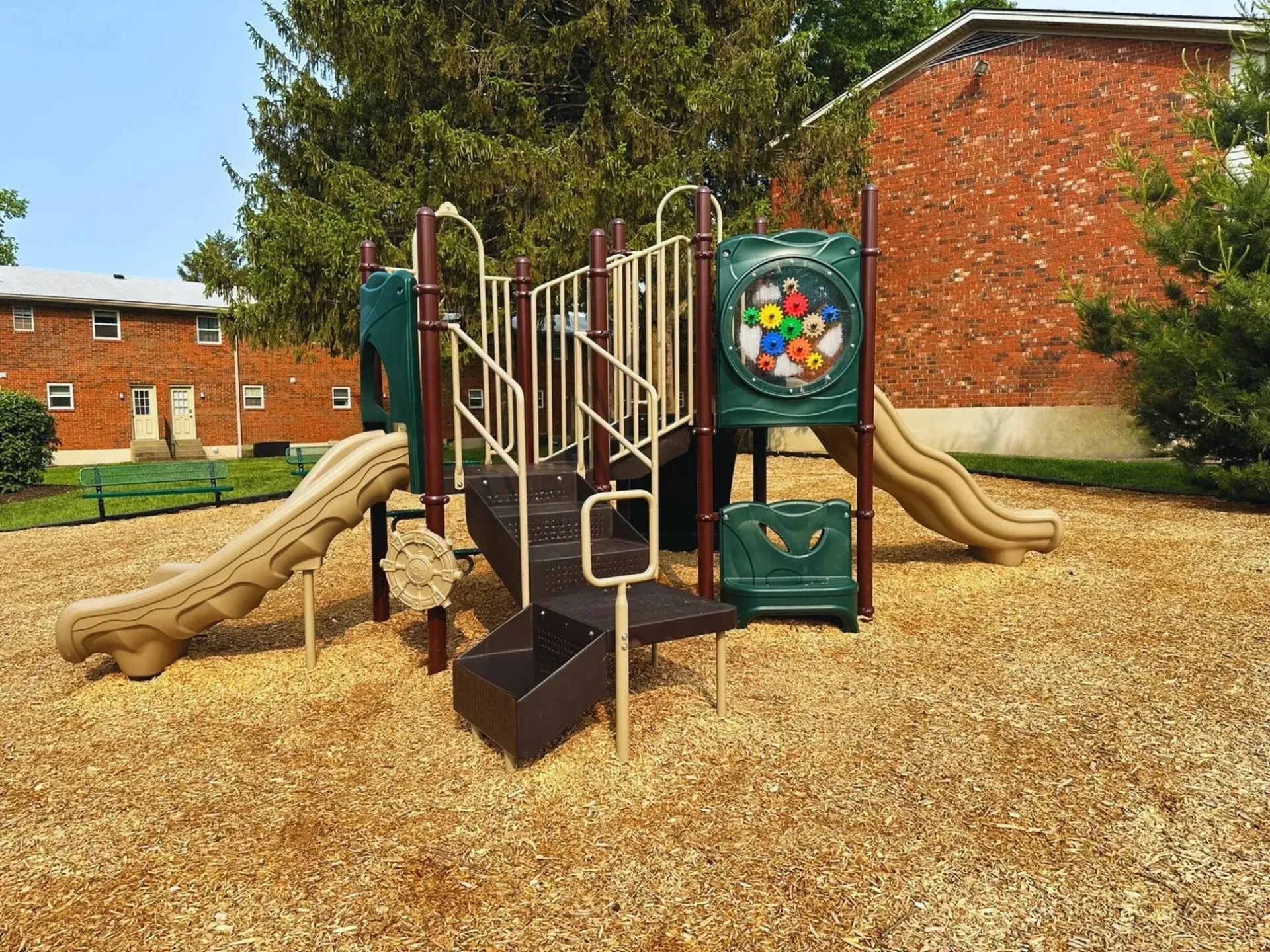 Playground with tan slides, brown wood chips, and red brick buildings in the background.