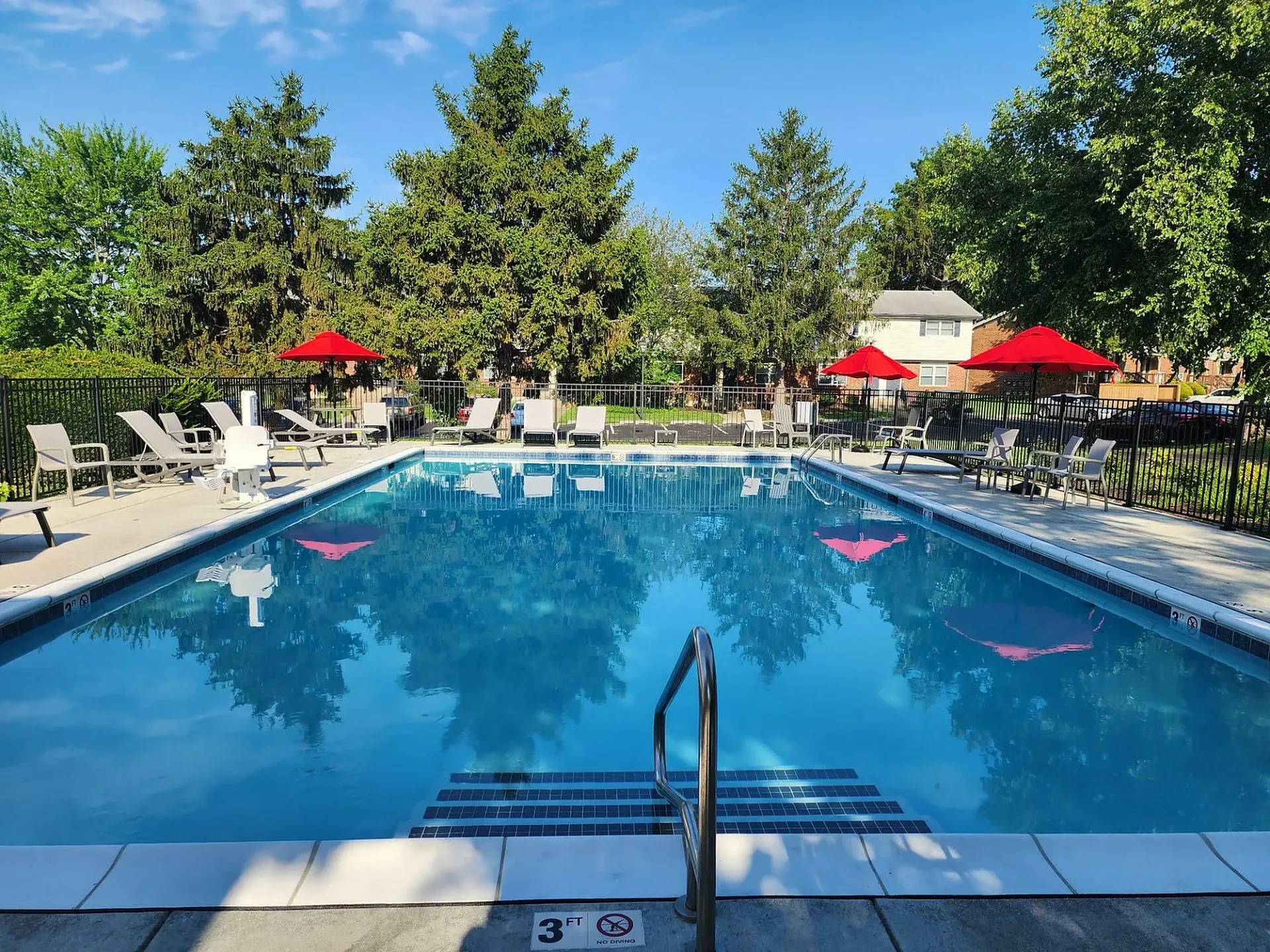 A rectangular pool with blue water surrounded by lounge chairs and red umbrellas, trees in the background.