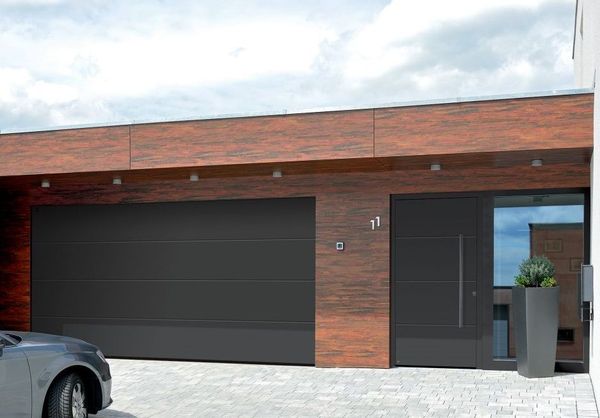 Portone grigio del garage di una casa bianca, fiancheggiato da pietre e piante. Vialetto d'accesso pavimentato.
