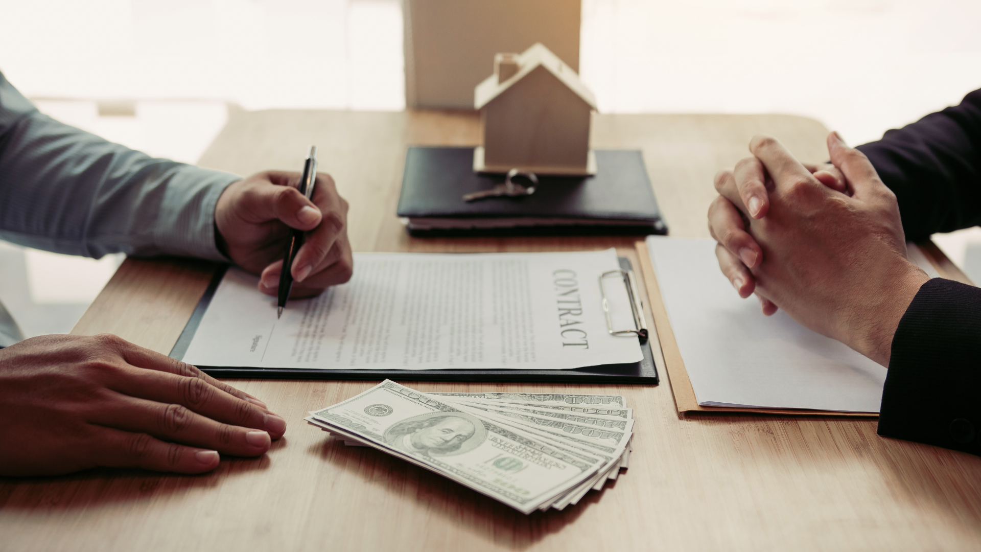 Hands signing mortgage papers across a desk with a small house model and cash in the center