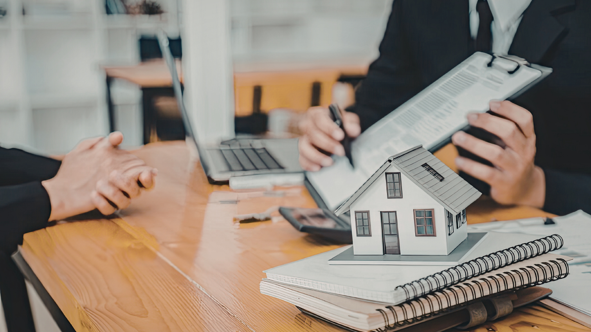 Hands discussing a house model and laptop at a desk, with paperwork and notebook in a real estate meeting
