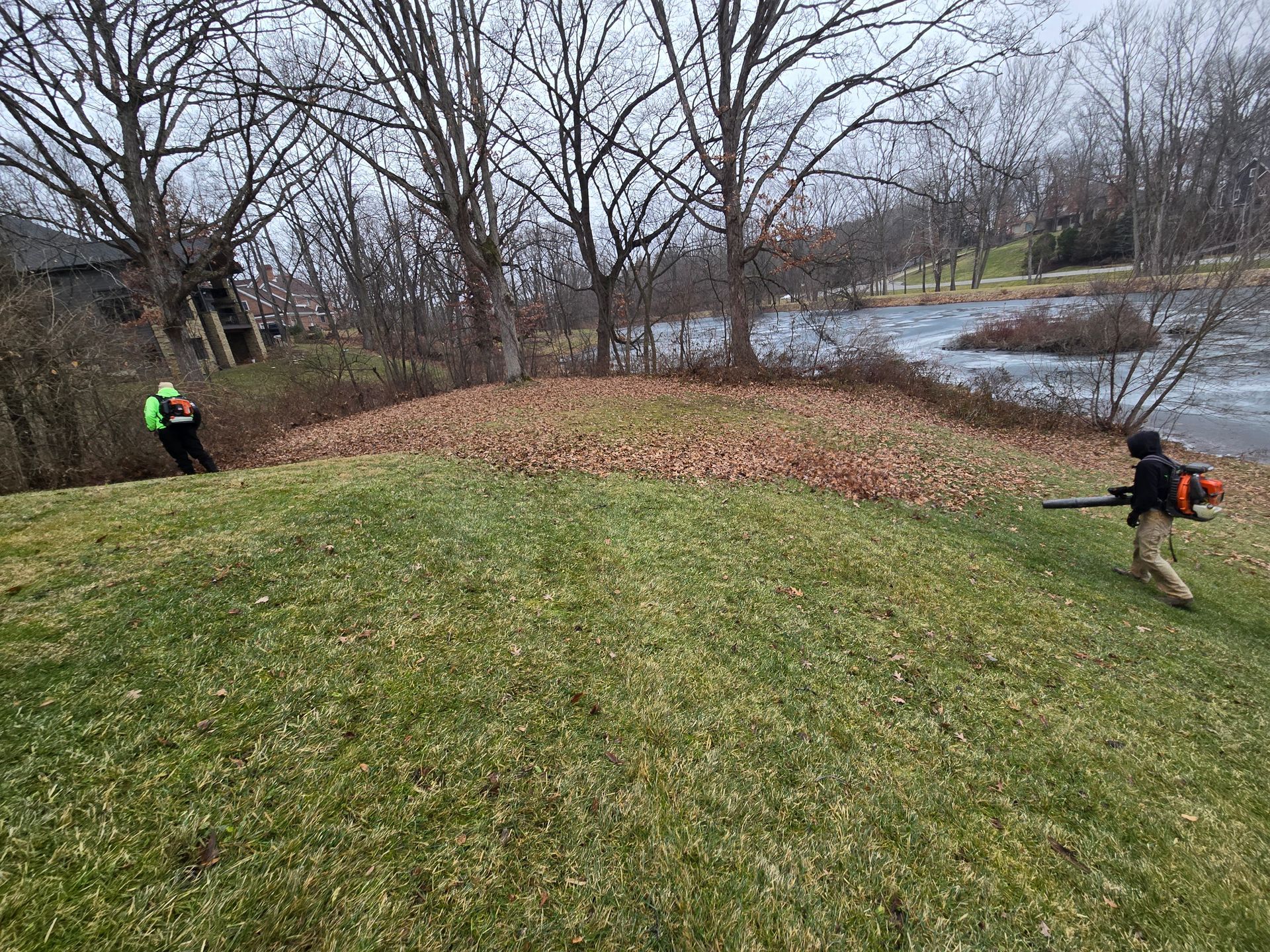 Two people in bright jackets work on a grassy riverbank with leafless trees in late fall.