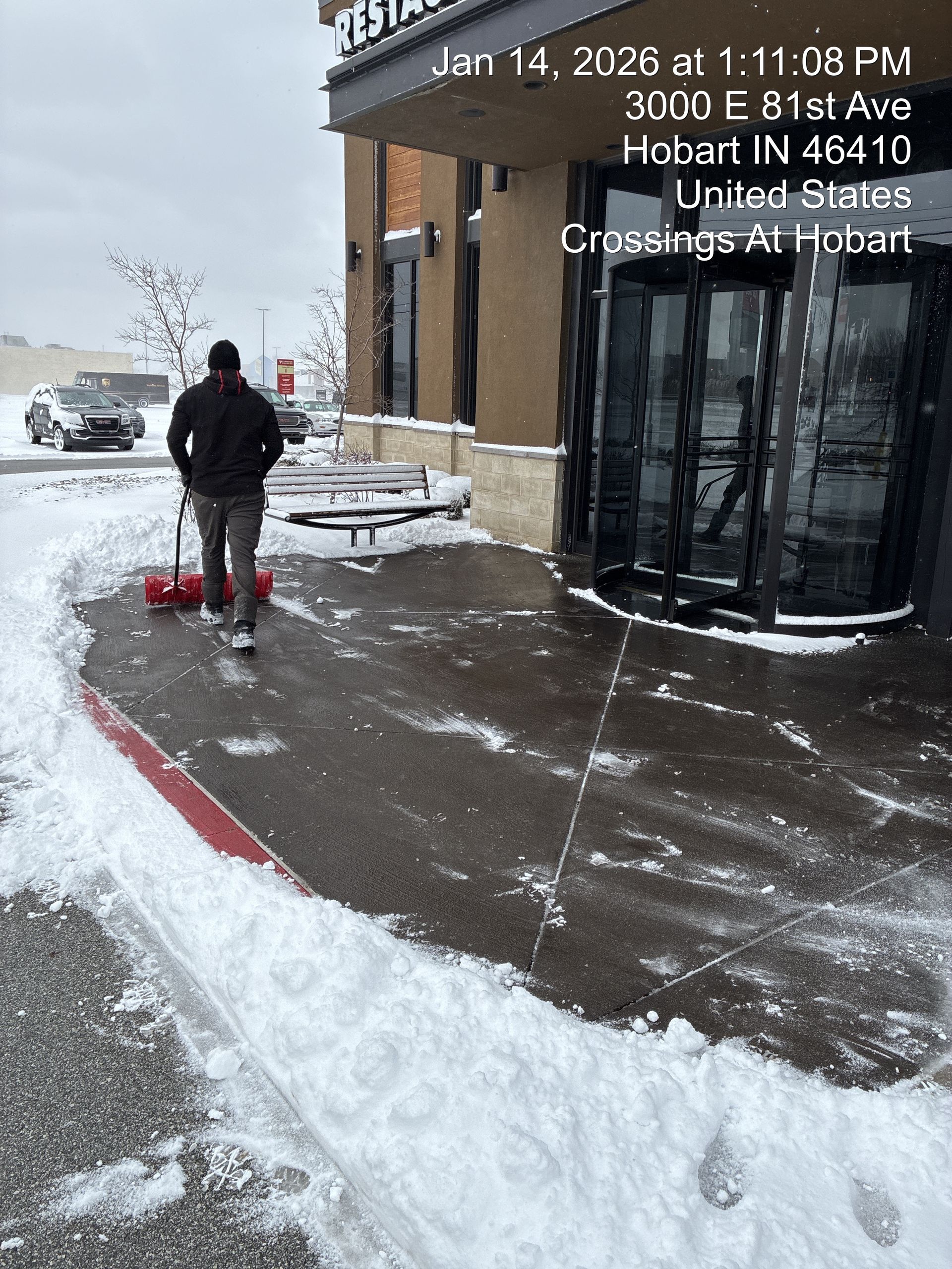 Person shoveling snow on a snowy sidewalk outside a building, with snow piled along the curb.