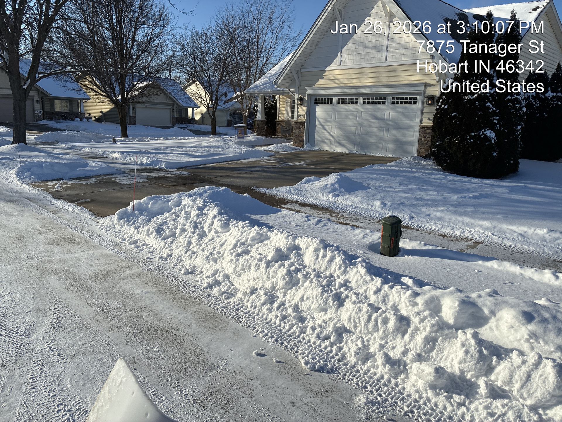 Snow-covered suburban street with a house, driveway, and piled plowed snow in front of bare trees.