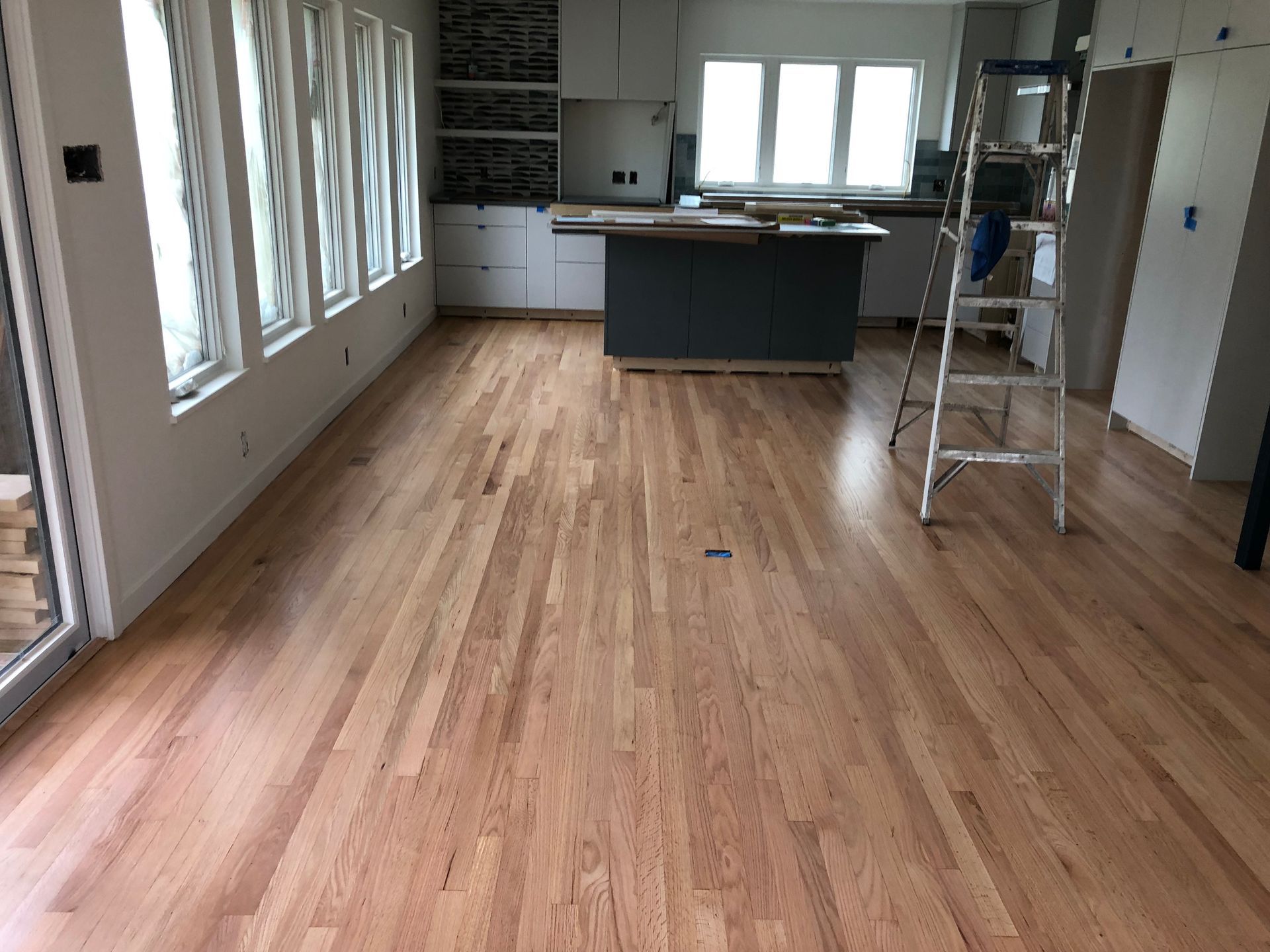 Hardwood floor in a kitchen area.  White cabinets, blue island, ladder, large windows, and an open doorway.