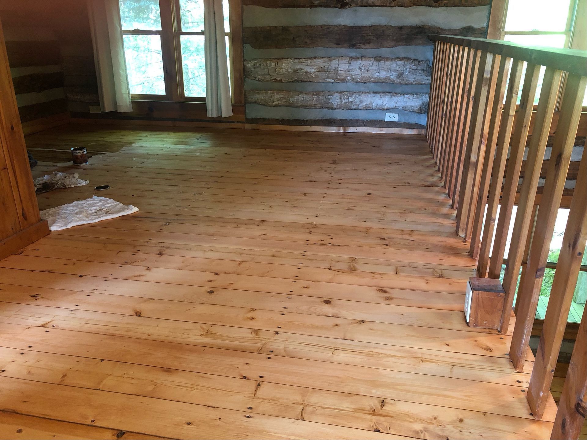 Refinishing wooden floor with handrail and log walls, near a window.