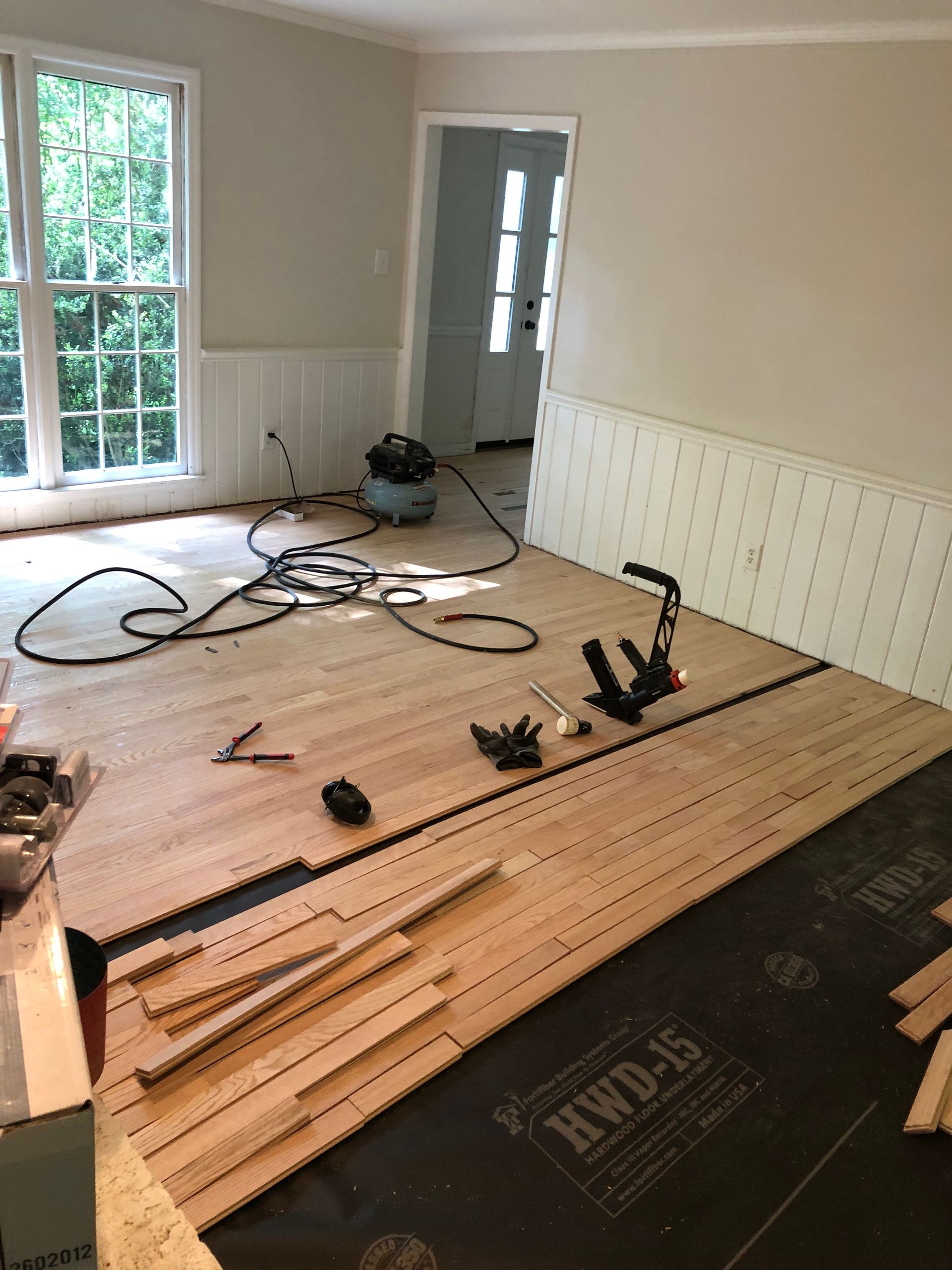 Hardwood flooring installation in a room with a window and white walls. Tools and wood planks are visible.