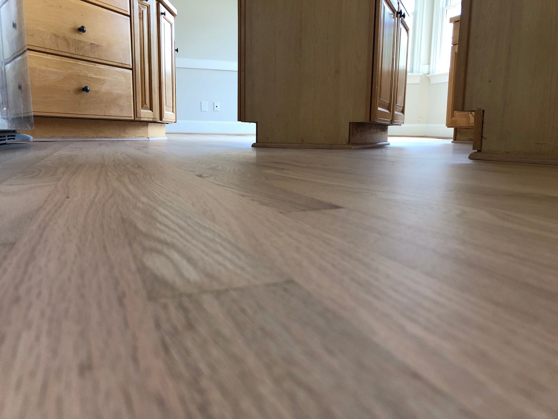 Low-angle view under wooden kitchen cabinets across a light wood floor toward a bright doorway