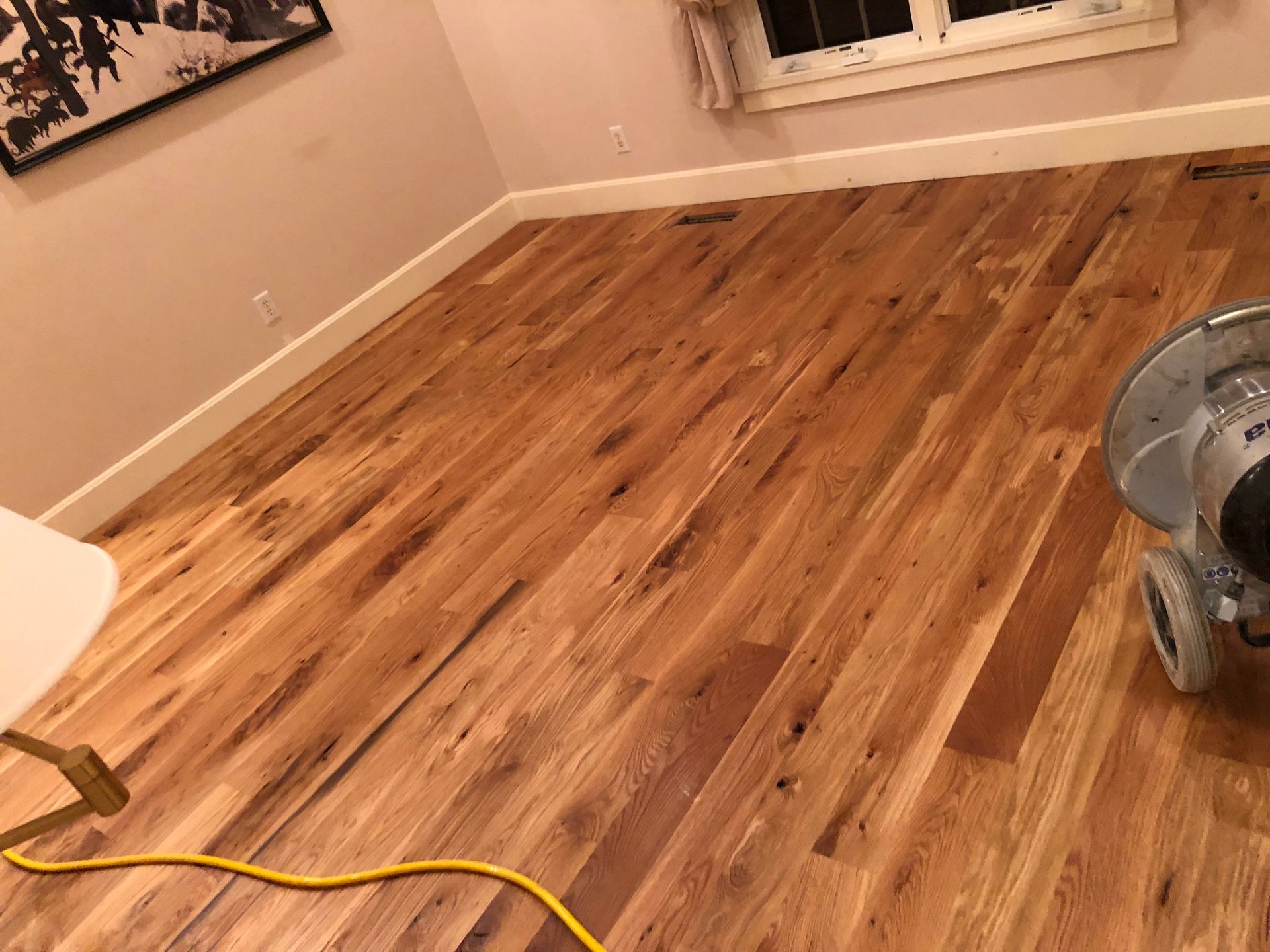 Hardwood floor being sanded; sander on right, white trim and wall, yellow cord.