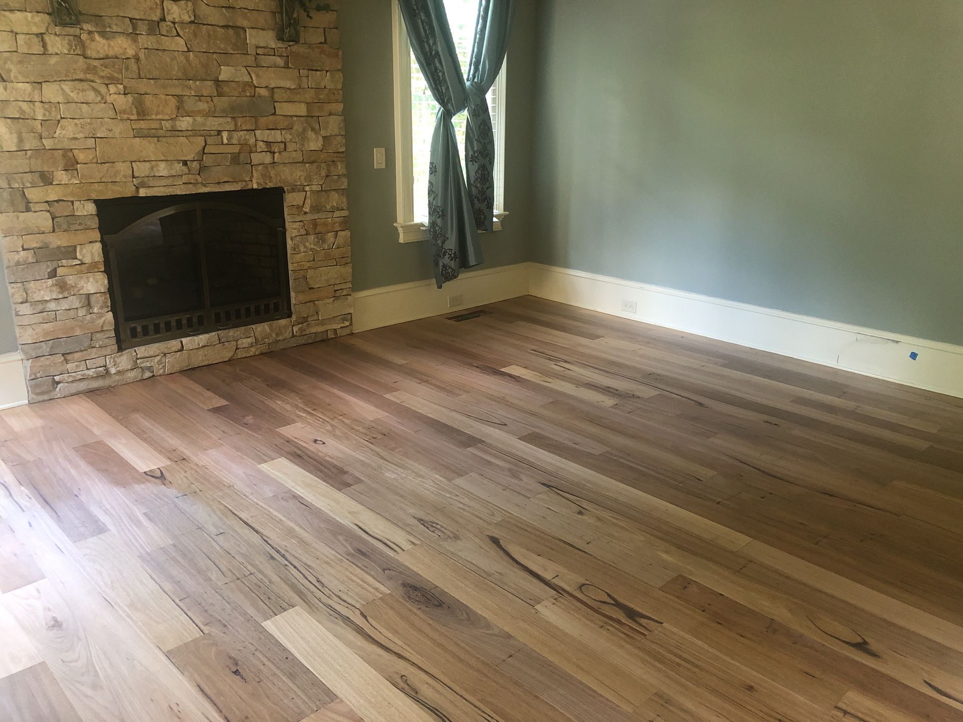 Hardwood floor in an empty room with a stone fireplace, and a window with curtain. Hardwood floor refinishing Raleigh NC.
