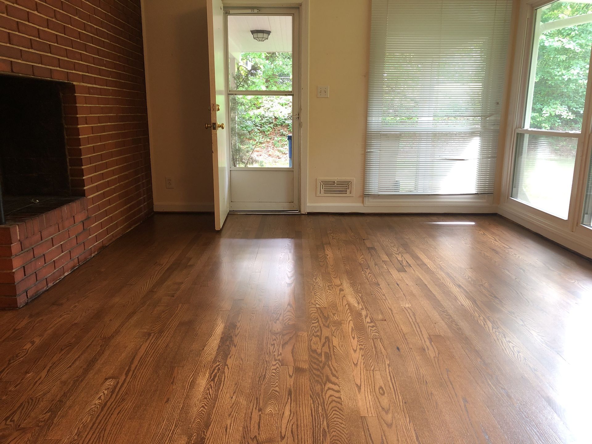 Hardwood floor in a room with a brick fireplace, open door, and windows with blinds.