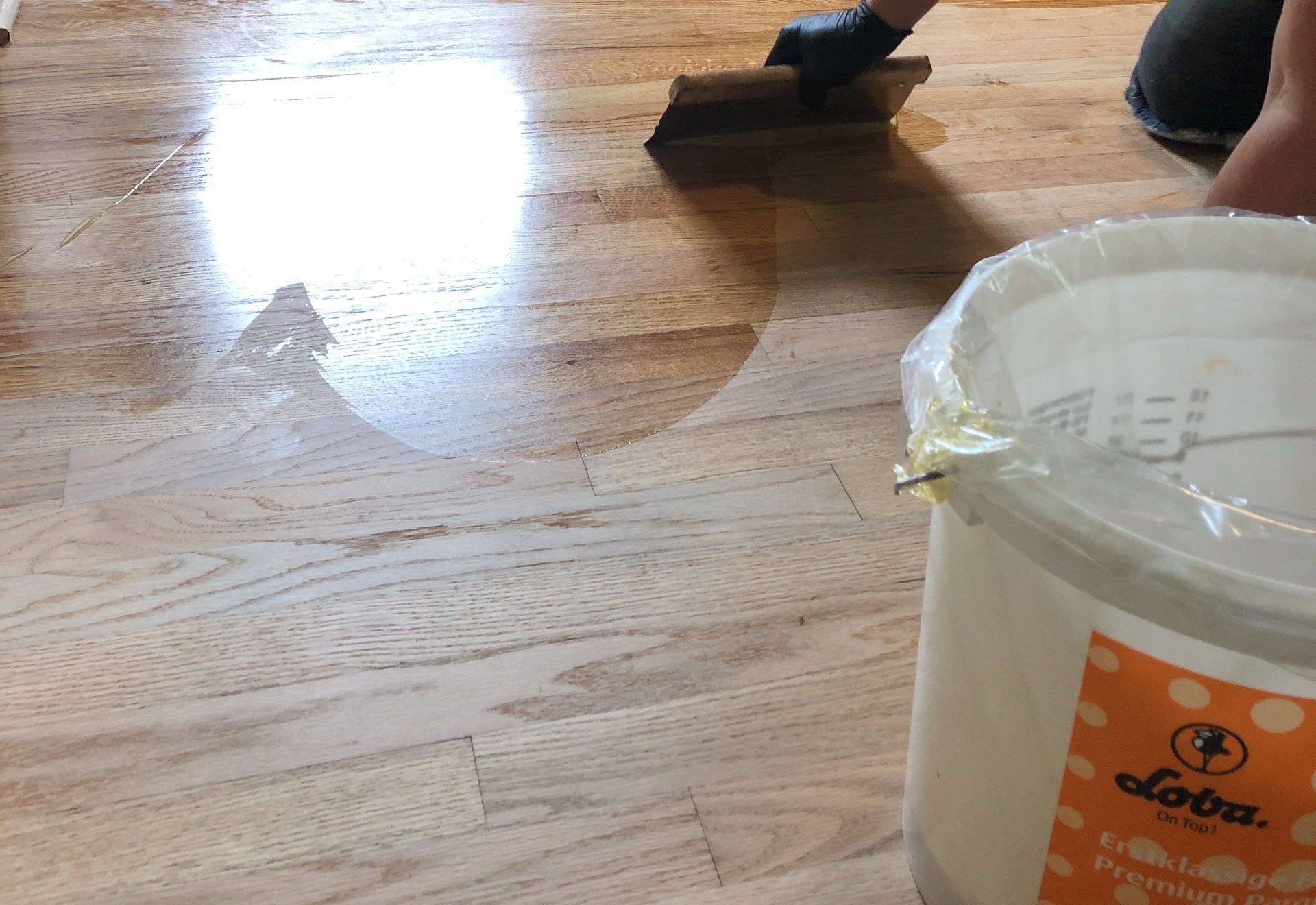 Person applying natural oil finish to hardwood floor with a trowel, bucket of Loba finish in foreground.