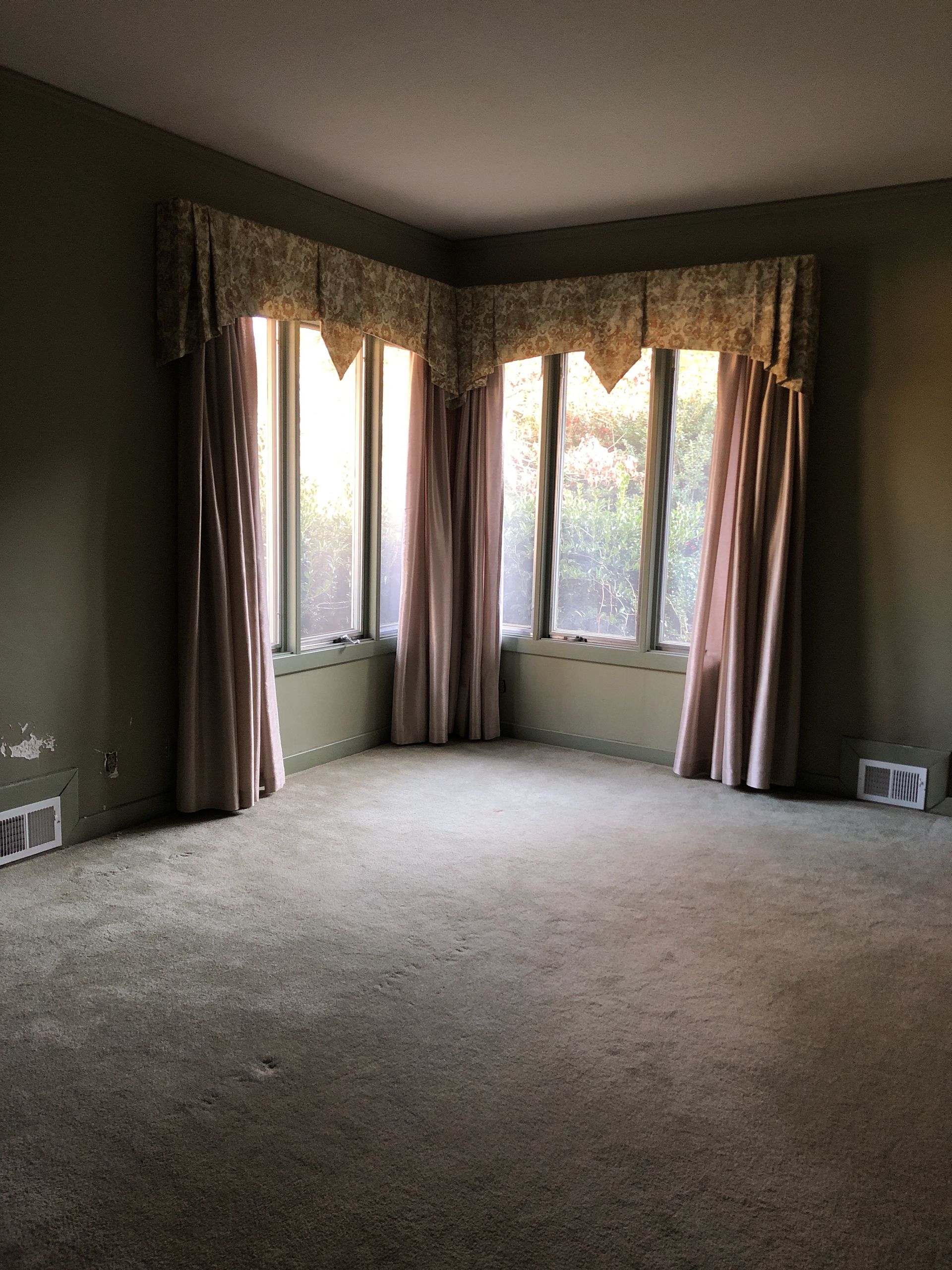 Empty room with a corner window. Green walls, beige curtains, and stained carpet.