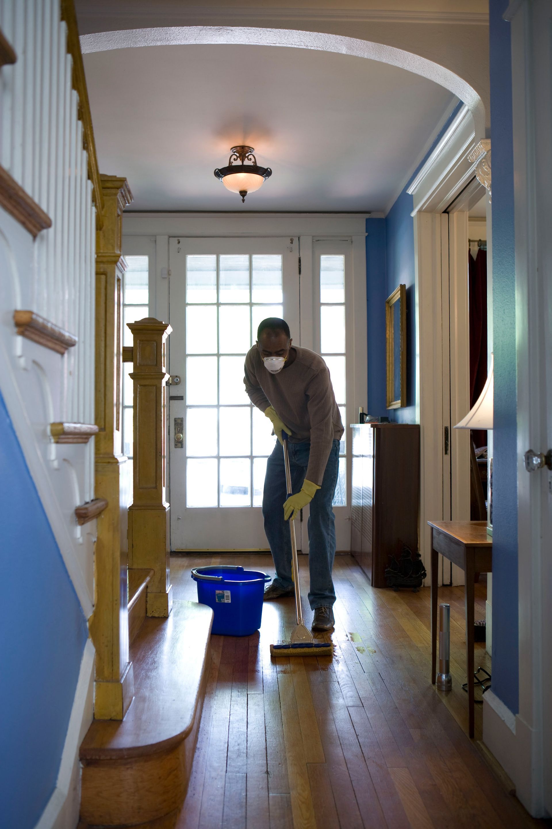 Person wearing face covering and gloves mopping hardwood floor in a hallway with stairs and a door in the background.