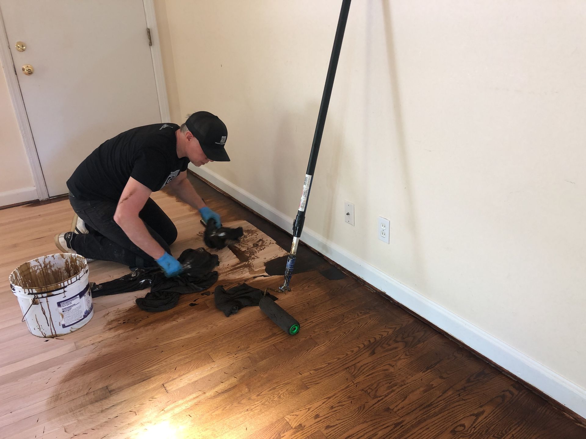 Person kneeling, staining hardwood floor with a roller. Brown stain, black shirt, blue gloves, white bucket.