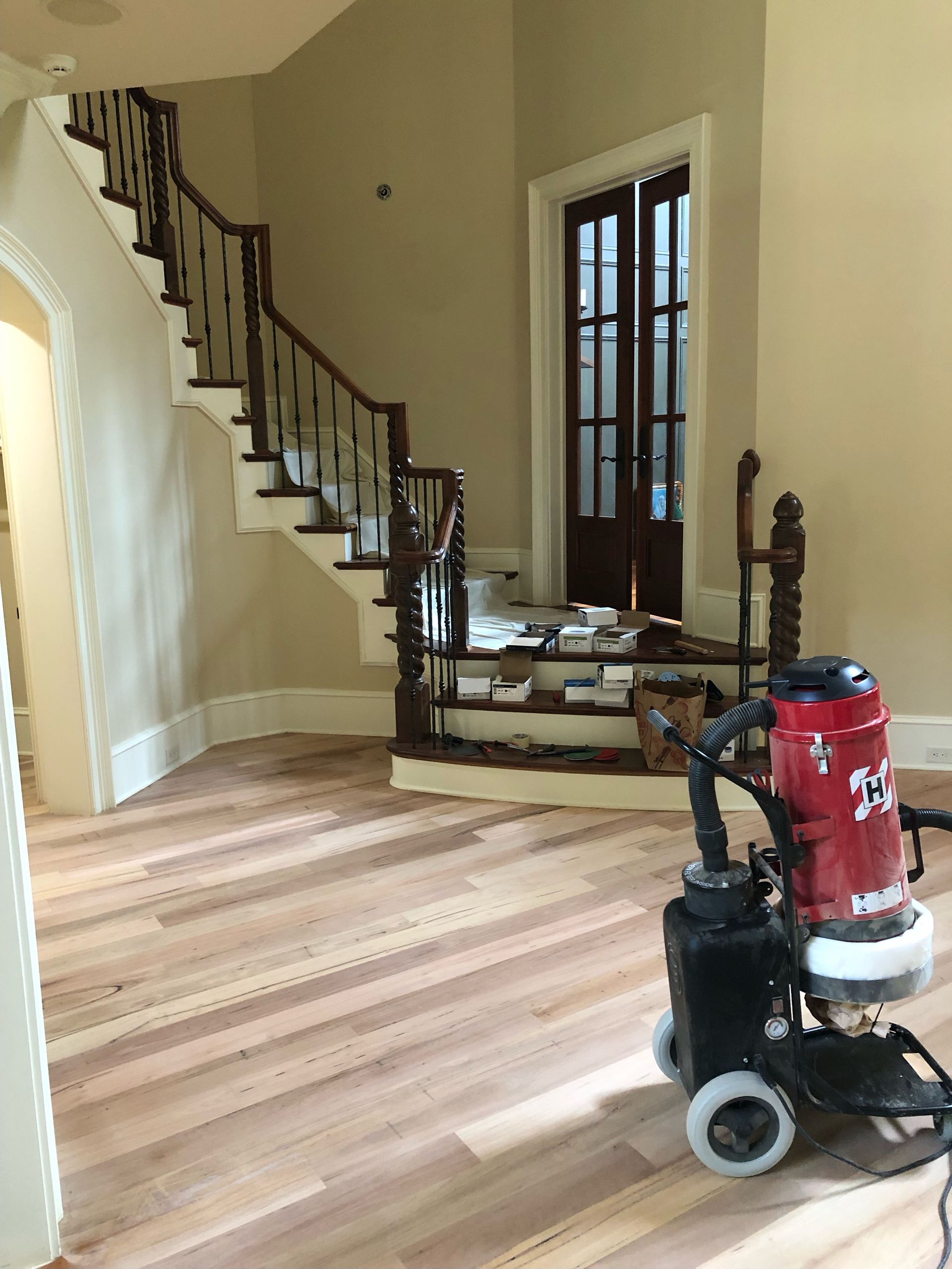 A dust vacuum is sitting on a freshly sanded wooden floor next to a staircase.