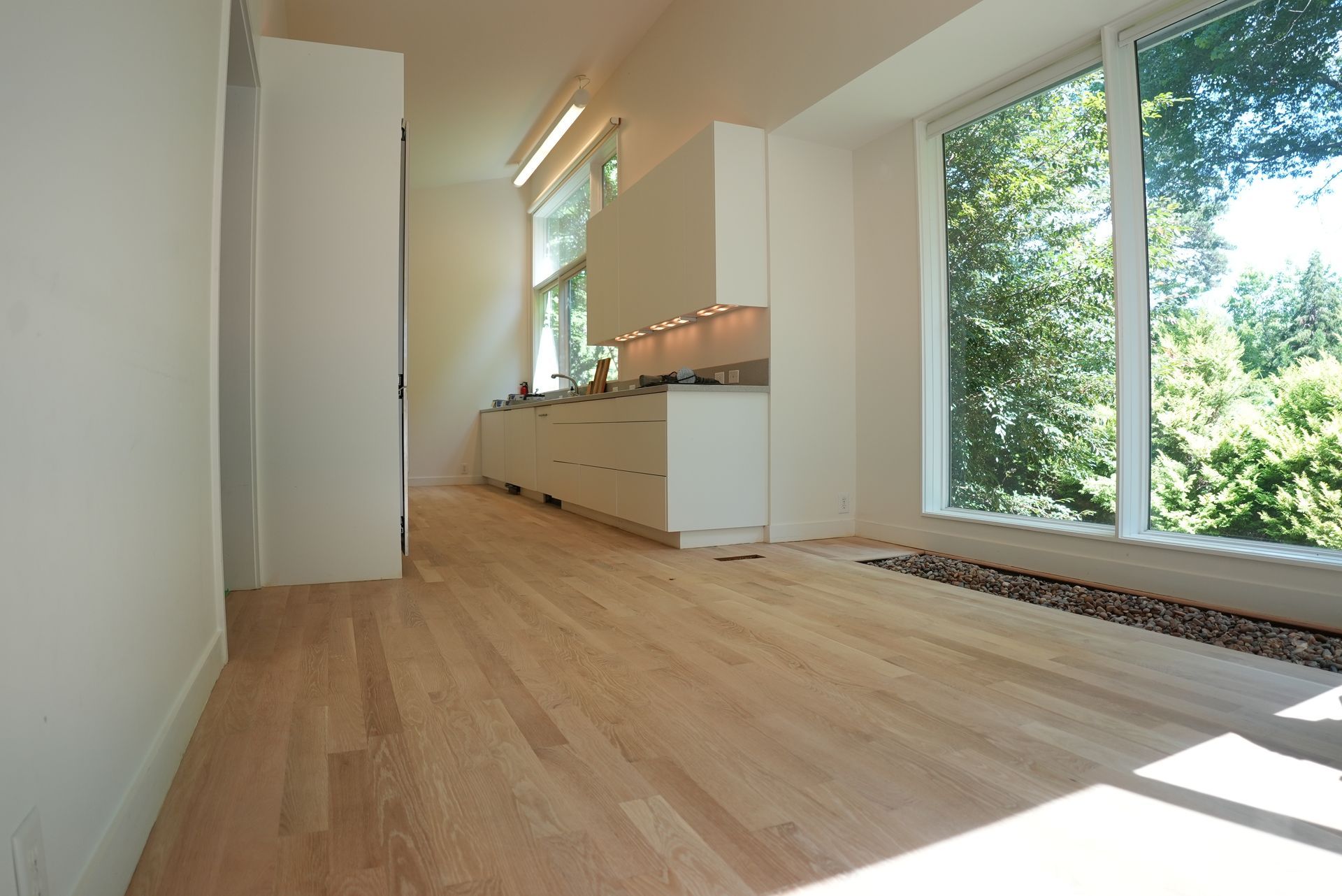 Light-filled room with hardwood floors, large windows overlooking lush greenery, and white cabinetry.