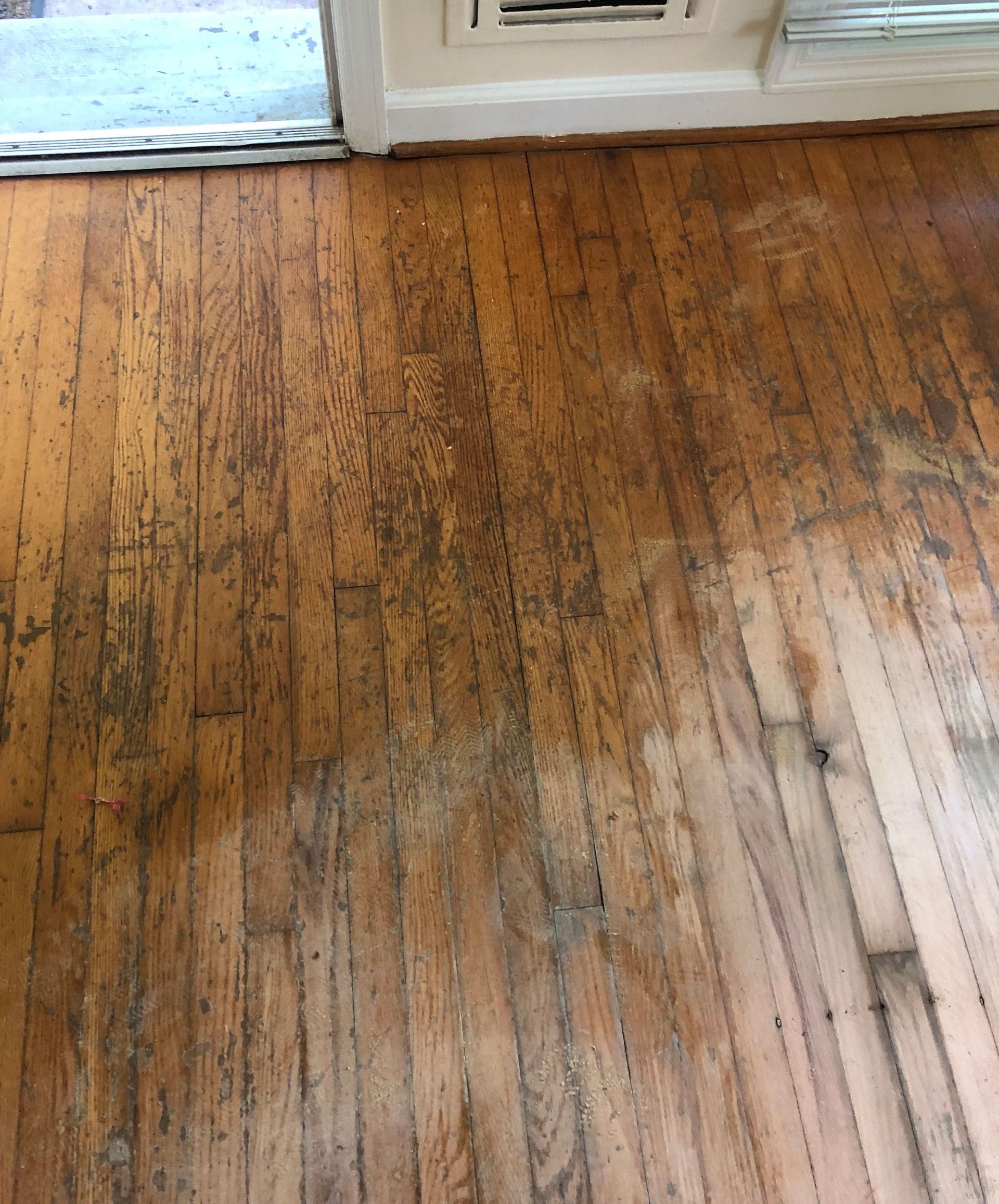 Worn hardwood floor with dark stains and sunlight near a window and baseboard.