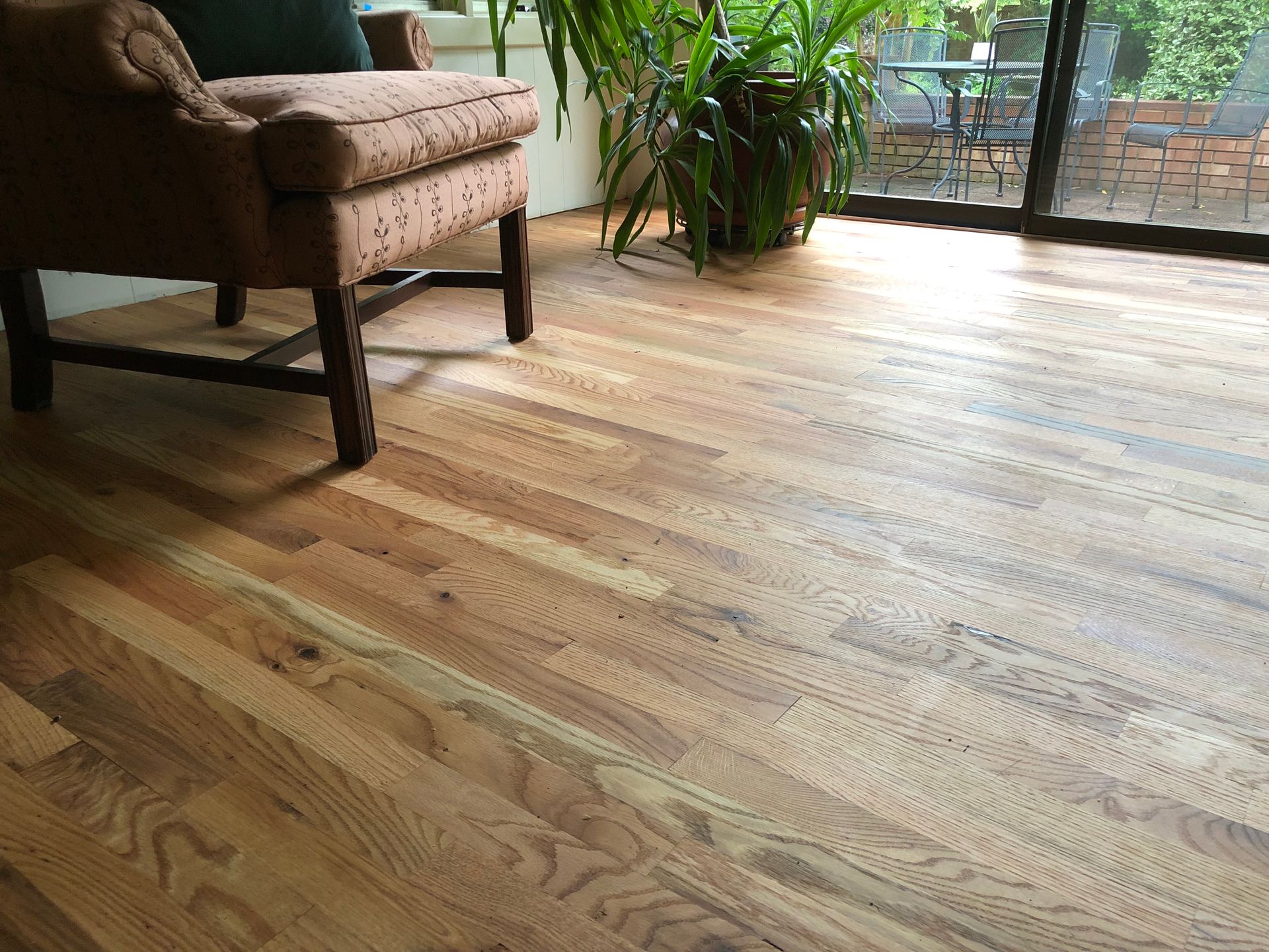 Wooden floor in a room with a brown armchair and a potted plant by a sliding glass door.