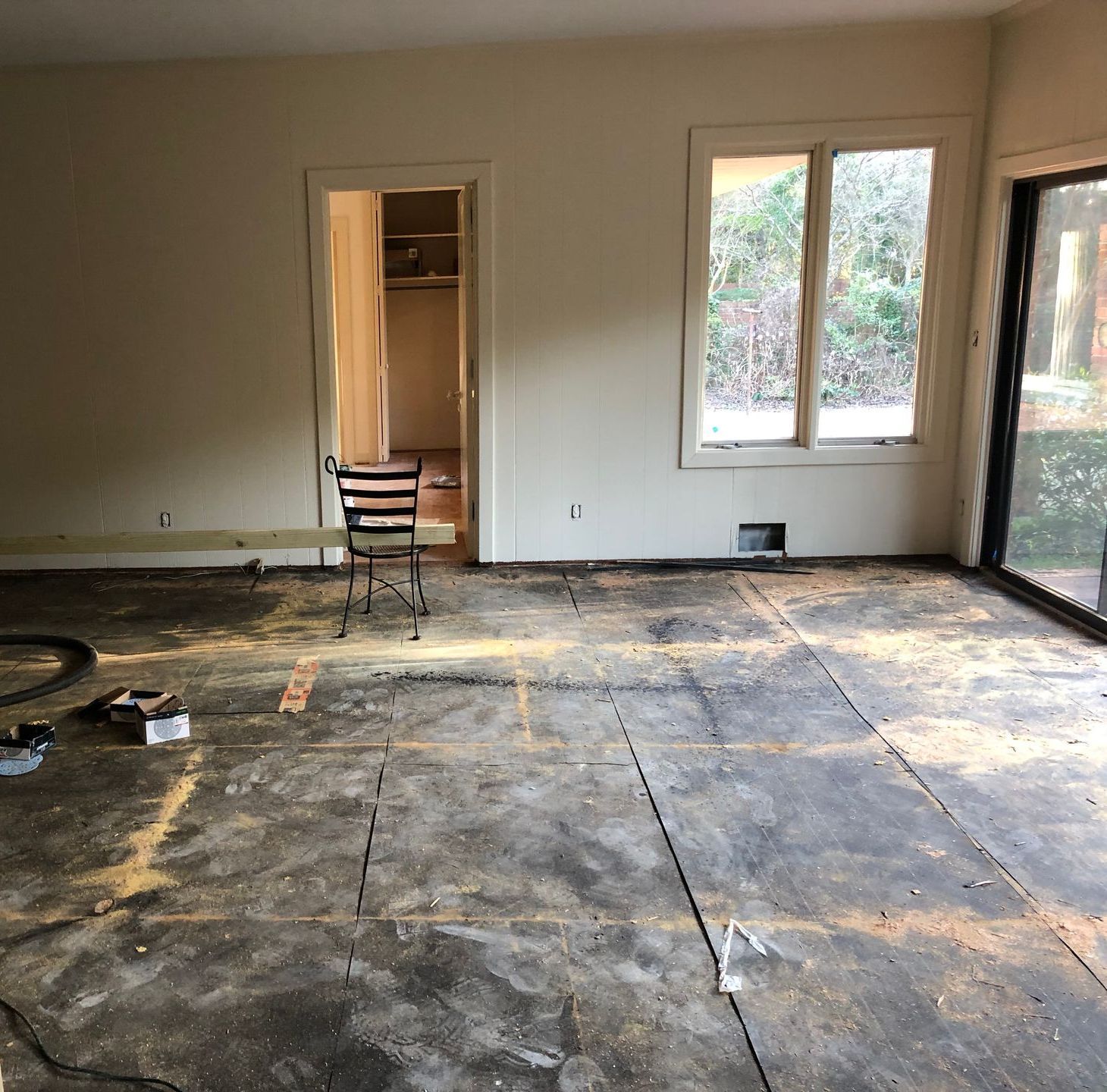Empty room with exposed floorboards; a chair sits in the middle. Walls are off-white; a window and sliding door are visible.