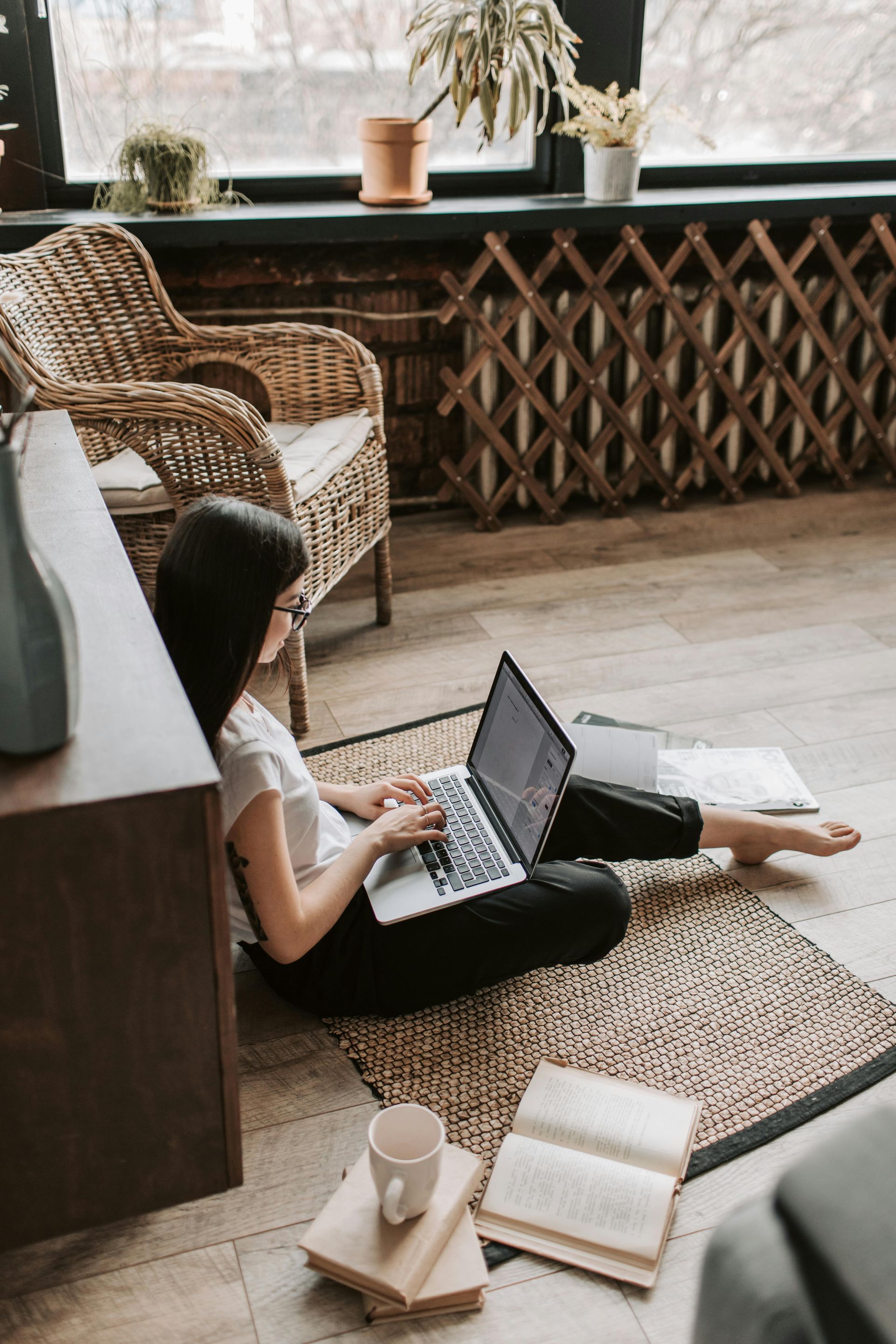 Woman sitting on floor with laptop, working near window with plants and wicker chair.