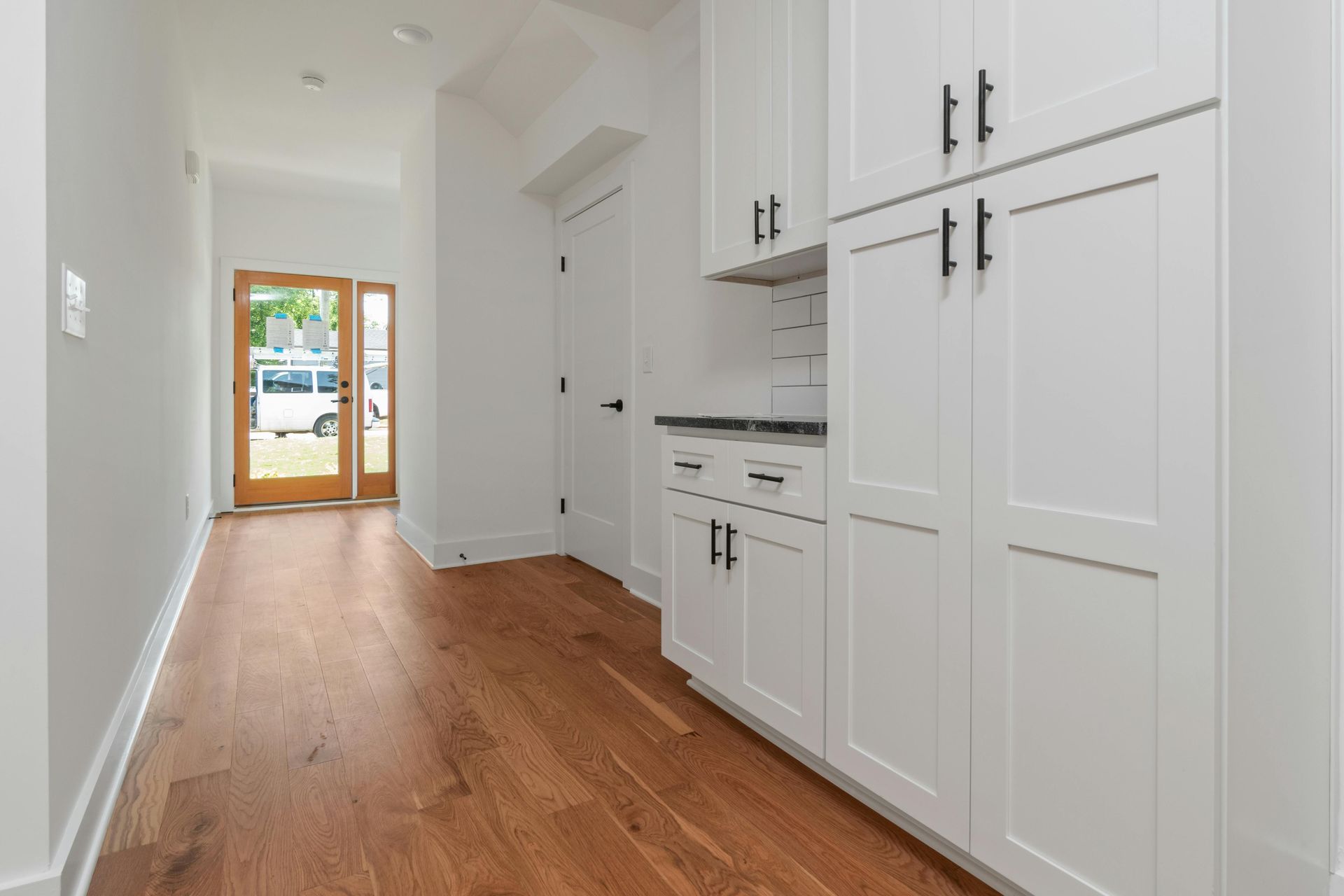 A narrow hallway with hardwood floors, white cabinets, and an open wooden door to the outside.